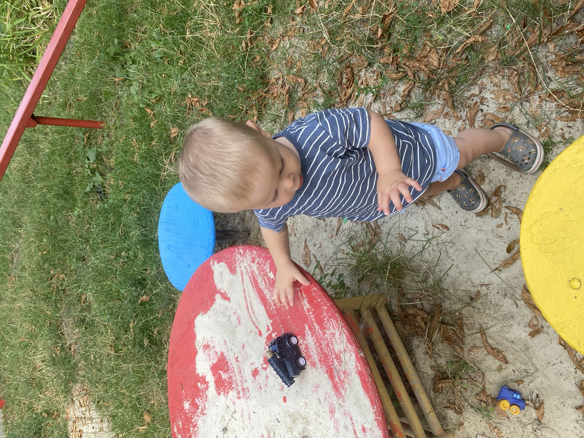 A blond-haired toddler wearing a blue and white striped t-shirt and blue shorts stands in a sandbox. The toddler is reaching out a hand towards a small black toy truck on a red and white circular table. A blue mushroom-shaped seat is visible behind the toddler. The sandbox is surrounded by grass and fallen leaves. A red metal bar is visible in the upper right corner. The toddler is wearing grey clogs with colorful designs. A small blue and yellow toy vehicle is on the sand near the base of the red and white table. Part of a yellow circular table is visible in the lower right corner.