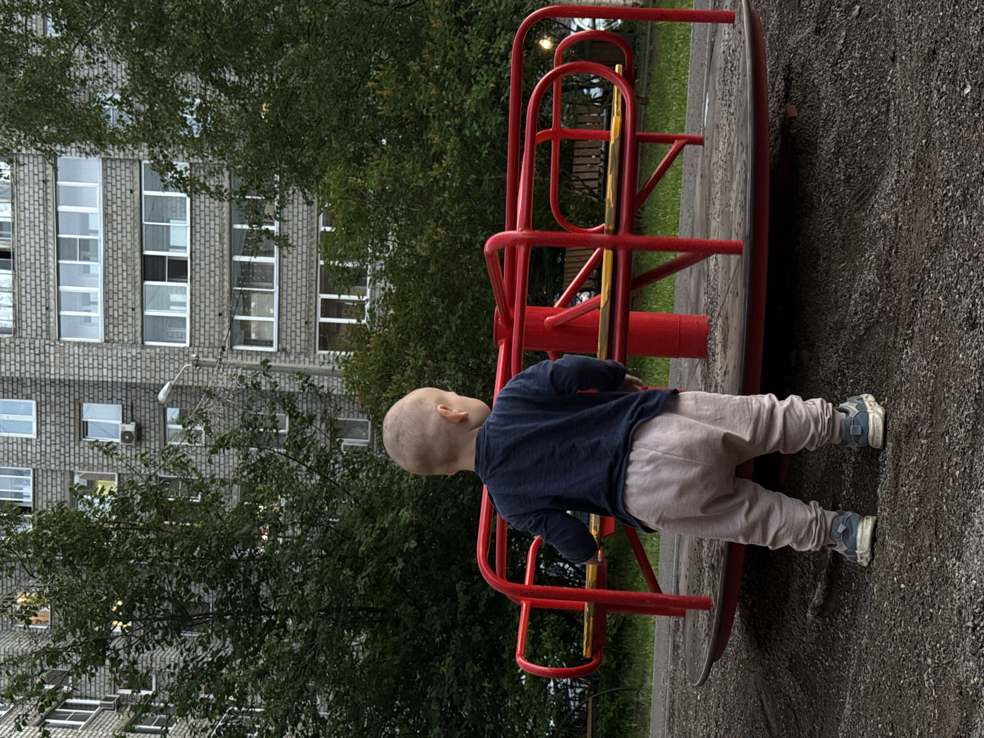 A young child, seen from behind, stands on a playground merry-go-round in Krasnoyarsk, Russia. The child is wearing a dark blue long-sleeved shirt and light pink pants, with gray sneakers. The merry-go-round is red with yellow seating. In the background, a grey brick apartment building with numerous windows is visible, partially obscured by lush green trees. The ground is covered in gravel and patches of grass.