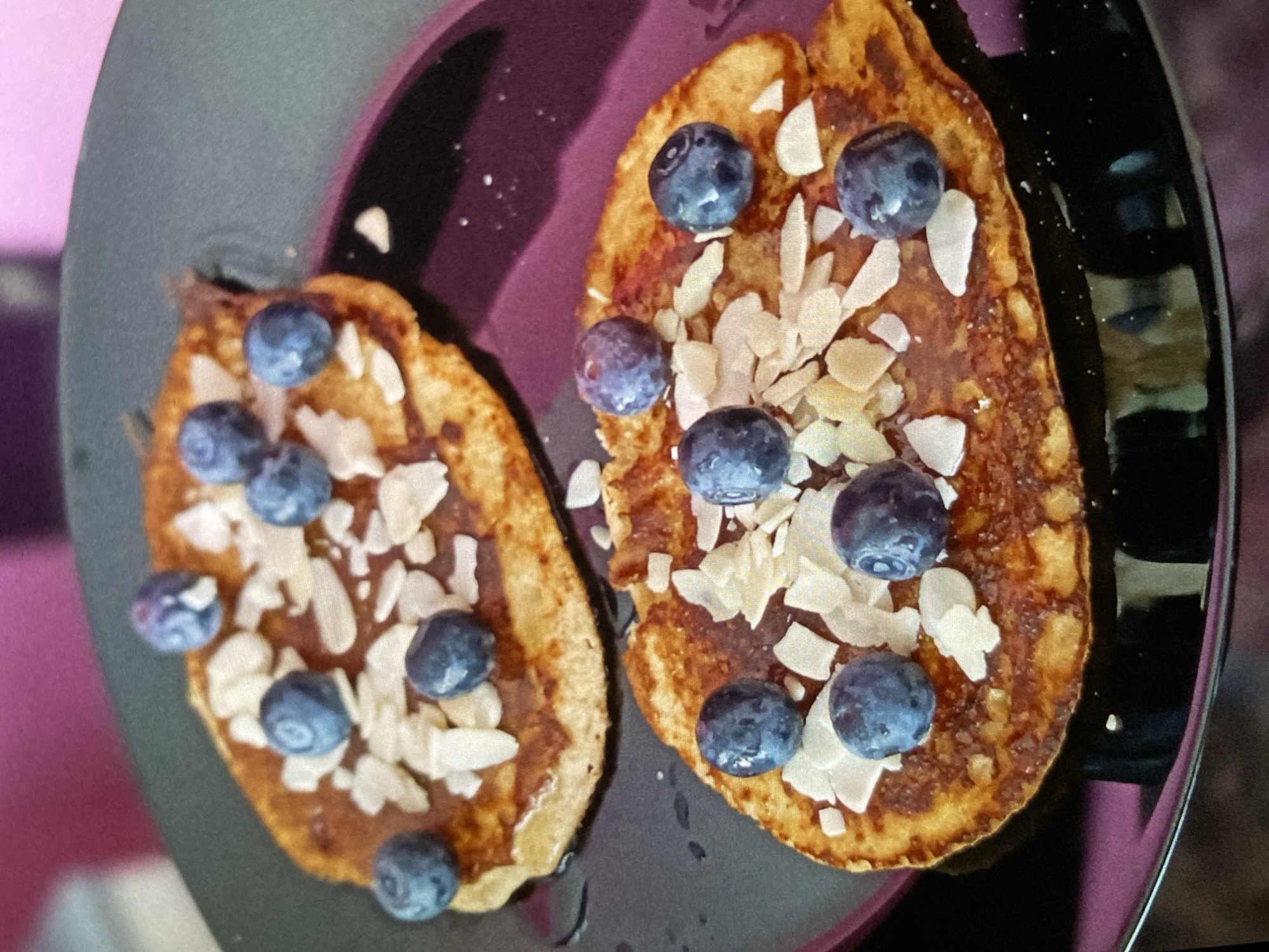 The media file displays a close-up, overhead view of two oval-shaped pancakes or fritters, generously garnished with fresh blueberries and slivered almonds. The food is presented on a dark, possibly non-stick, cooking pan or serving platter.
The pancakes are golden-brown, indicating they are well-cooked, and appear to have a light drizzle of sauce, possibly syrup or chocolate, on their surface. Numerous plump, deep blue blueberries are scattered across both pancakes, along with delicate white slivered almonds, adding both texture and visual appeal. The dark surface of the pan or platter provides a strong contrast, making the colorful toppings stand out. Subtle reflections are visible on the glossy, raised edge of the pan on the right side.
The background is softly blurred, showing hues of pink and dark purple, suggesting an indoor setting, likely a kitchen or dining area. There are no people visible in the image. The scene captures a ready-to-eat meal, presumably breakfast or brunch, prepared with attention to presentation. No visible text is present in the image. The location context for this scene is Bankya, Bulgaria.