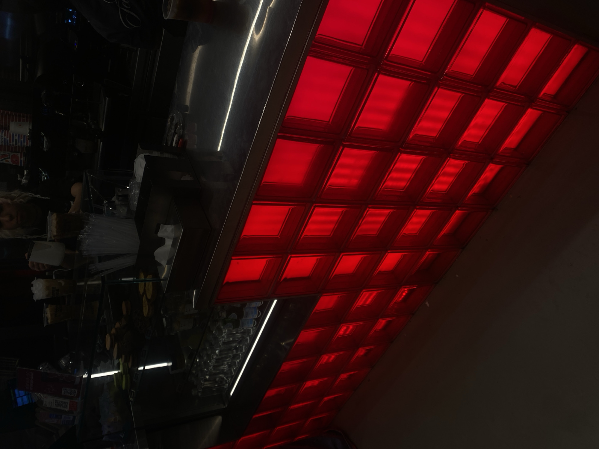 The image shows a commercial establishment in Yerevan, Armenia. A counter with an illuminated, red-tiled front is prominent. Behind the counter, food items, drinks, and disposable utensils are visible, suggesting a cafe or food service environment. The lighting is dim, and reflections create a sense of depth within the confined space.