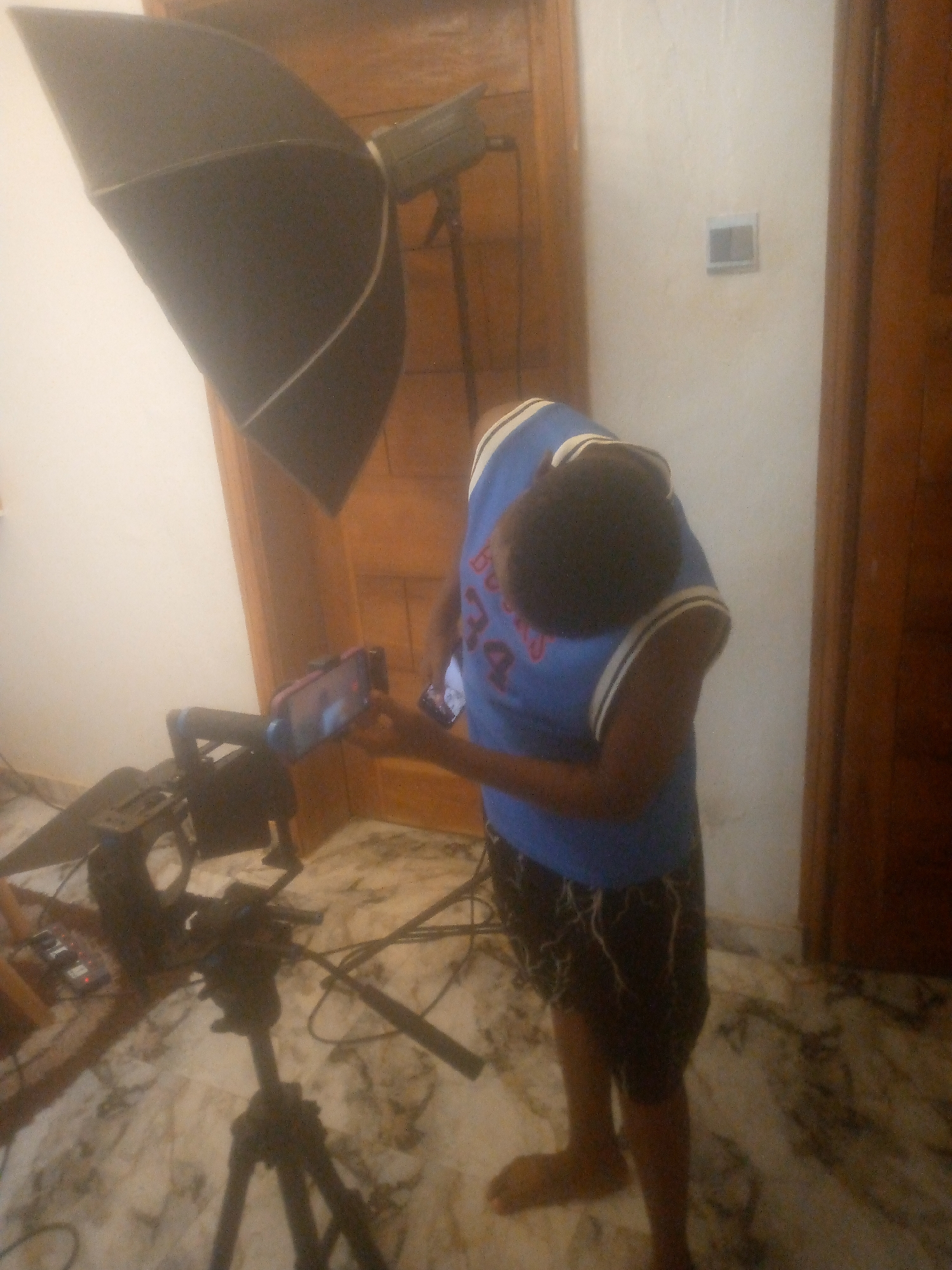 A person in a blue and white basketball jersey and black shorts is operating a smartphone mounted on a tripod. A large black umbrella light is positioned to the left of the camera. The scene appears to be indoors, possibly a home studio, with a light switch on the wall and a brown door. The floor is marble. The specific location is Ikorodu, Nigeria, but there are no unique markers in this image that can confirm this.