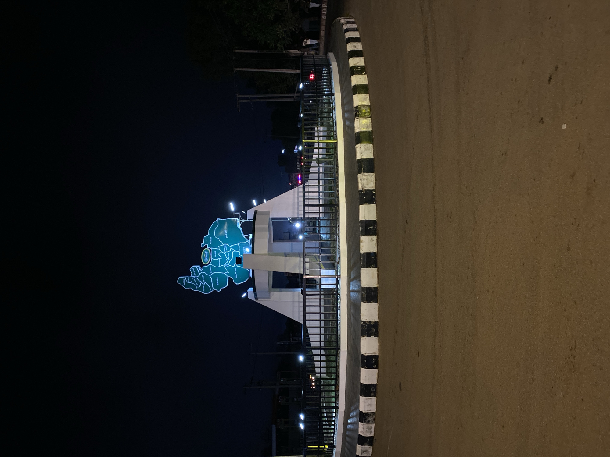 The image shows a night scene at a roundabout in Kuru Dungus, Nigeria. A prominent monument in the center of the roundabout features a large, illuminated map of a region, with several place names visible. The roundabout itself is surrounded by a black metal fence and has a curb painted with alternating black and white stripes. The road surface appears to be asphalt. Several streetlights illuminate the scene, casting a warm glow. In the background, there are trees, utility poles, and some distant buildings. A few figures, possibly pedestrians, can be seen on the right side of the image. The overall atmosphere is that of a well-lit urban space at night.