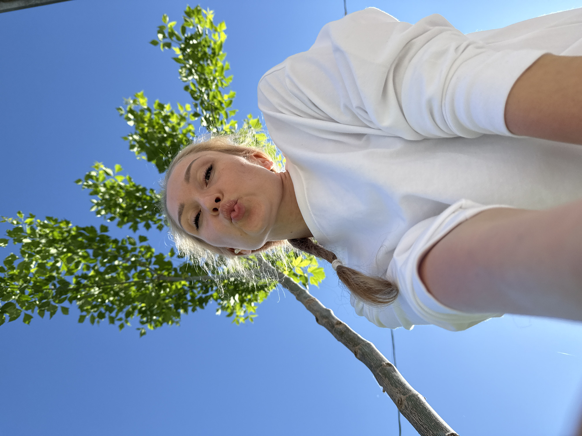 A low-angle selfie of a fair-skinned woman with blonde hair in a braid, wearing a white long-sleeved shirt, is set against a bright blue sky and a small tree with green leaves. The woman is puckering her lips towards the camera. The tree trunk is thin and light brown. The location is likely outdoors, with the blue sky suggesting a clear day. It is not possible to determine if the image was taken in Chaikovsky, Russia, based on visual evidence alone.