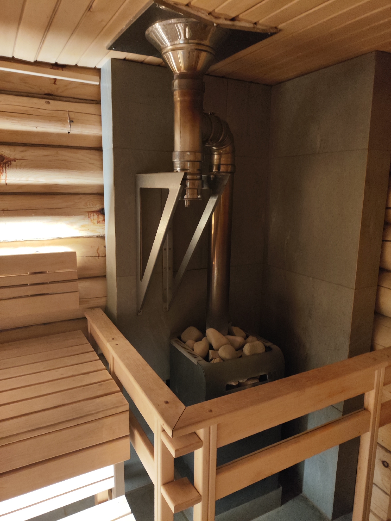 The image depicts the interior of a traditional Russian banya (sauna) in the village of Staroye Martyánovo, Russia. The scene is dominated by natural wood construction, with log walls and a slatted wooden ceiling. In the center of the image, a stainless steel sauna stove is visible, filled with stones. A metal chimney system extends upwards from the stove and connects to an opening in the ceiling. To the left, wooden benches are arranged, providing seating within the sauna. The overall impression is one of rustic simplicity and traditional design, typical of a rural Russian sauna. There are no people or animals present in the image.