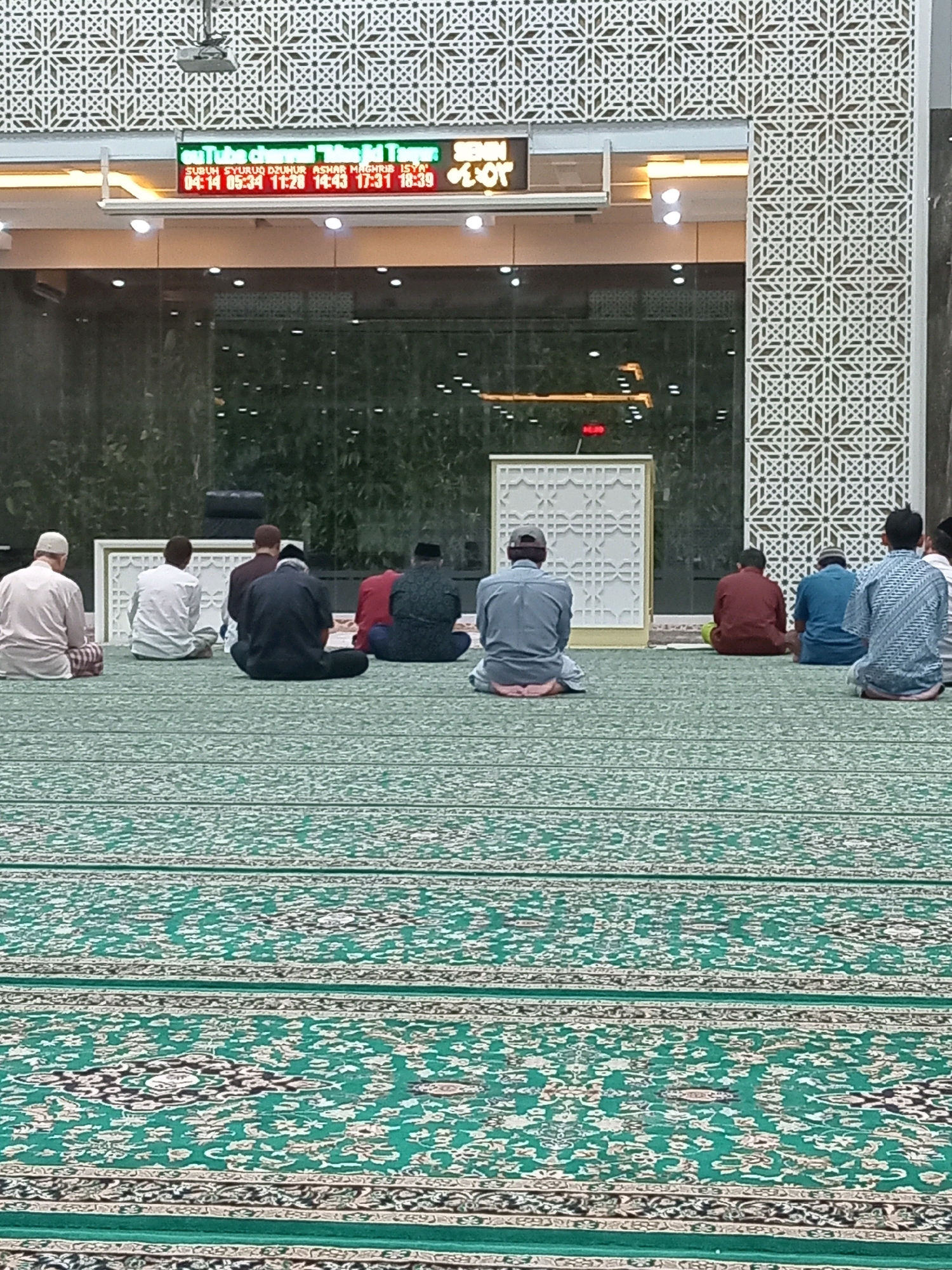 The image shows the interior of a mosque, specifically the prayer hall, with a green carpet covering the floor. Several men are seen from behind, seated on the carpet in prayer positions, facing a decorative wall. In the background, a digital display shows prayer times and what appears to be a channel name, "Tube channel TIKTOKTAYAR". The mosque's architecture includes geometric patterns on the walls and pillars, and a large glass window in the background reveals trees and greenery outside, suggesting an outdoor view. The overall scene depicts a religious gathering during prayer time in a mosque. The presence of the digital display with prayer times further reinforces the context of a place of worship. The city of Randuagung, Indonesia is mentioned as the location, indicating the specific geographic setting for this mosque.