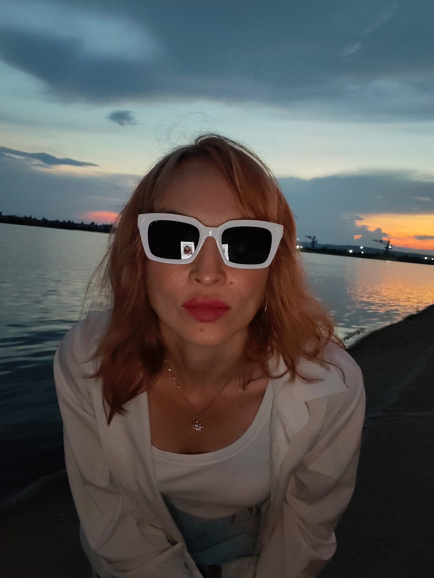 A woman with auburn hair is seated outdoors at dusk, looking directly at the camera. She is wearing large white-framed sunglasses, a white top, and a white unbuttoned shirt. A small, circular pendant necklace is visible on her chest. The sunglasses reflect a small image of what appears to be a person. Behind her, a body of water stretches towards the horizon, where a colorful sunset is visible. The sky transitions from deep blue to orange and pink hues. In the distance, along the horizon, several tall industrial structures, possibly cranes, are silhouetted against the sky. The area to the right of the woman appears to be a paved or rocky embankment. The location is identified as the Kama River embankment in Chaikovsky, Russia.