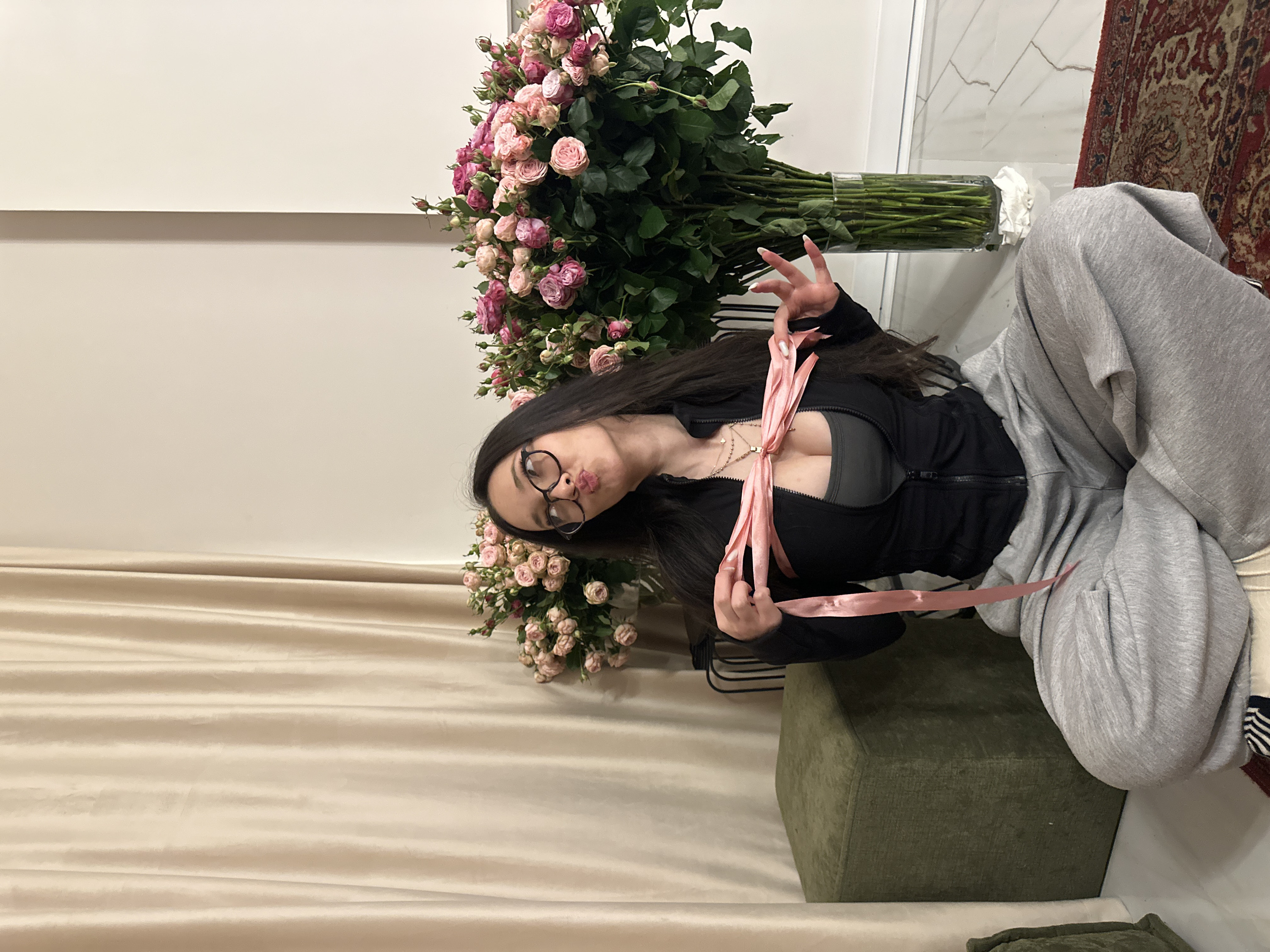 The image captures a young woman posing playfully in an indoor setting, surrounded by a large bouquet of flowers. The photograph is oriented vertically, with the woman positioned on the right side of the frame.
The central figure is a woman with long, dark hair that cascades around her shoulders. She is wearing round, dark-framed glasses and has a pouting expression on her lips, possibly indicating a playful or cute mood. Her nails are long and shaped, featuring a light-colored, possibly French manicure style. She is dressed in a black long-sleeved top, which appears to be a zipped jacket, worn over a grey undergarment or top. She also wears loose-fitting grey sweatpants. A delicate gold-colored necklace with small pendants is visible on her chest.
She is holding and interacting with a long, light pink satin ribbon that appears to be loosely tied or arranged around her upper torso, creating a knot or bow-like detail at her chest. Her hands are positioned to hold the ribbon, with her fingers slightly curled.
To the woman's left, a voluminous bouquet of pink and light pink roses, possibly spray roses or miniature roses, dominates a significant portion of the scene. The flowers are arranged in a tall, clear glass vase, through which the green stems are clearly visible. The vase is placed on a light-colored, reflective surface, which could be a modern table or a polished floor.
The woman is seated on a dark green, upholstered cube-shaped ottoman or low stool. Further to the right, a portion of a traditional patterned rug with red, cream, and darker accents is visible on a light-colored, possibly tiled, floor with subtle grey veining.
The background consists of a light, off-white or cream-colored wall, which has a subtle horizontal line or shelf dividing it. Below this, there is draped, light beige or cream-colored fabric, likely curtains or soft furnishings. The overall lighting is bright and even, suggesting an indoor environment during the day or under artificial light. The image has a light, airy aesthetic, contrasting with the rich colors of the flowers and the woman's dark hair and clothing.