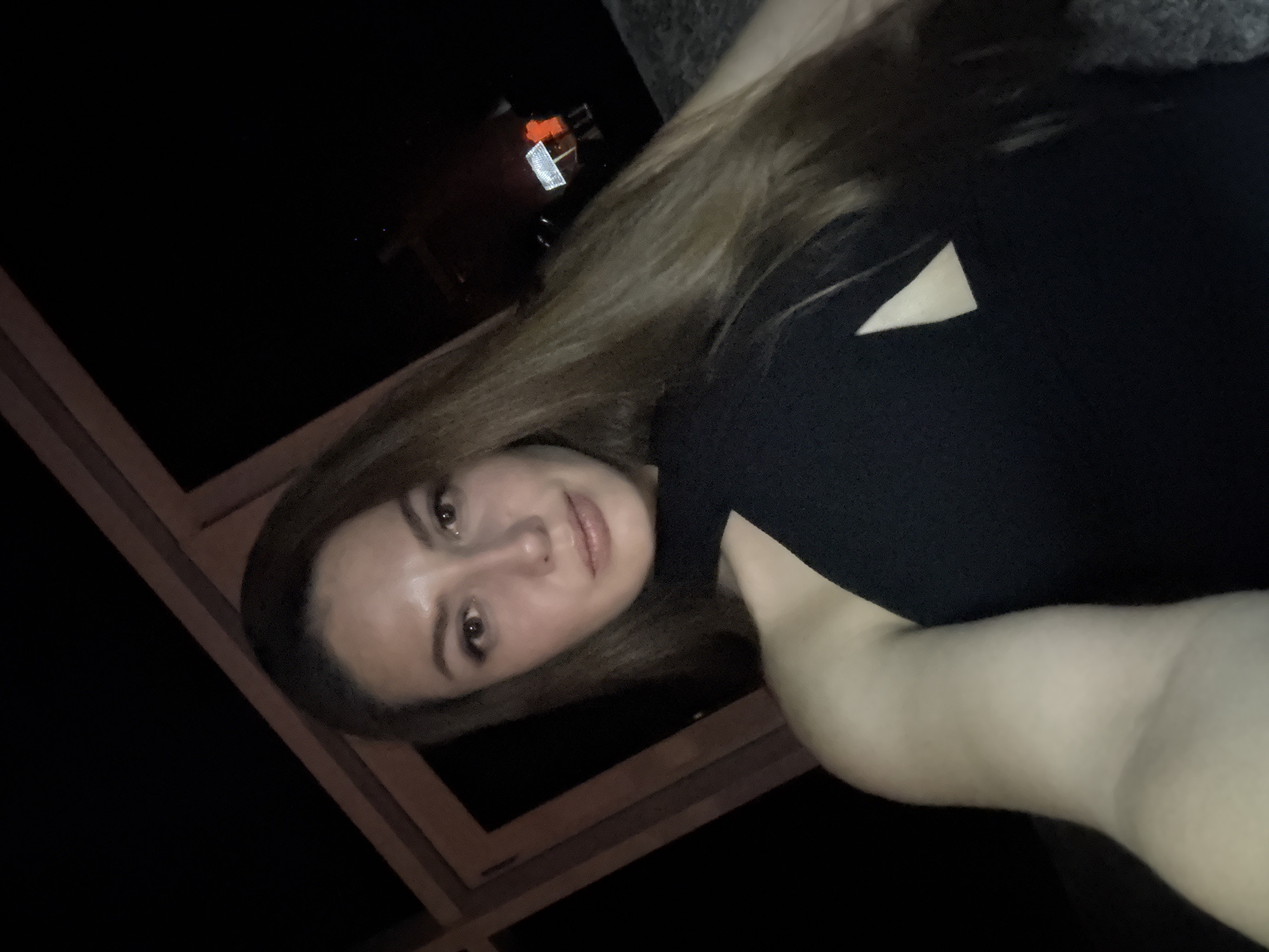 The media file is a low-angle, vertically oriented selfie of a young woman indoors at night. She has long, light brown hair and is wearing a black top with a small triangular cutout detail on the shoulder. Her face is turned towards the camera, and she has light makeup on, including eyeliner and lipstick. She appears to be looking slightly to her right with a neutral expression.
The background is predominantly dark, suggesting it is nighttime. In the upper left portion of the image, there is a hint of an indoor setting with what appears to be some furniture and possibly a distant light source, perhaps from a window or a reflection. A dark, angular object, possibly part of a window frame or furniture, is visible in the lower left and bottom of the frame. The lighting is dim, with some highlights on the woman's face and arm, indicating an artificial light source from the front or side.
There is no visible text in the image. The overall mood is casual and personal, typical of a selfie taken in a private space.