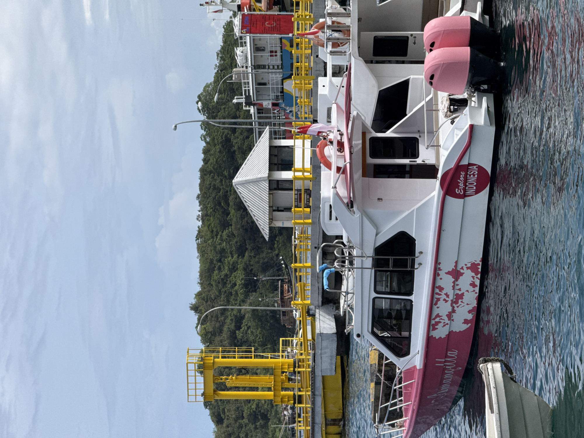 A vertically oriented, medium-distance outdoor shot shows a boat docked at a pier in Labuan Bajo, Indonesia. The boat is mostly white with red trim, featuring a red circle with the words "Explore INDONESIA" printed on it. The boat is docked at a yellow pier, alongside other boats. In the background, a building with a white roof is visible, as is a hill covered in green trees. The sky is blue with some white clouds. The water reflects the colors of the boat and sky.