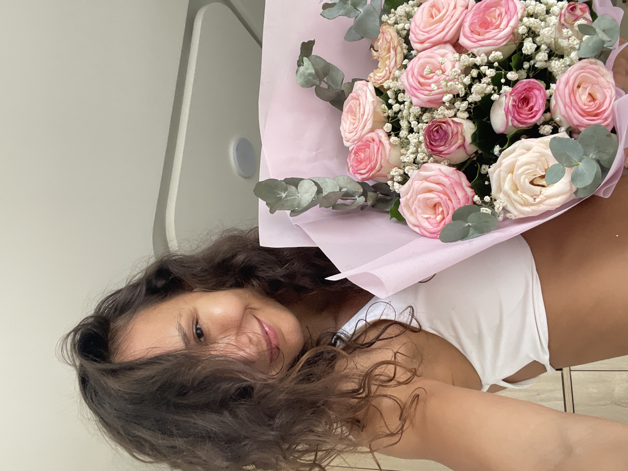 This is a vertically oriented, eye-level, medium close-up selfie taken indoors. The subject is a young woman with long, dark, curly hair, who is wearing a white top. She is holding a bouquet of pink and white roses with baby's breath and eucalyptus leaves wrapped in pink paper. Her face is partially obscured by her hair, but she is smiling. The background appears to be an indoor setting with a pale, textured wall and a thin, light-colored vertical element. The lighting suggests daytime. There is no visible text. The location context is Mũi Né, Vietnam, though there are no explicit visual cues in the image to confirm this.