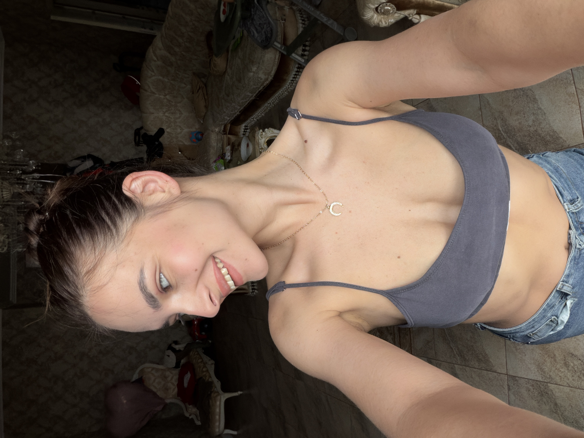 A young woman with her hair pulled up in a bun is shown in a close-up, shoulder-up shot. She is smiling and looking to her left. She is wearing a dark gray bra and blue jeans. She has a necklace with a crescent moon pendant. The background is a dimly lit room with ornate furniture and a patterned wallpaper.