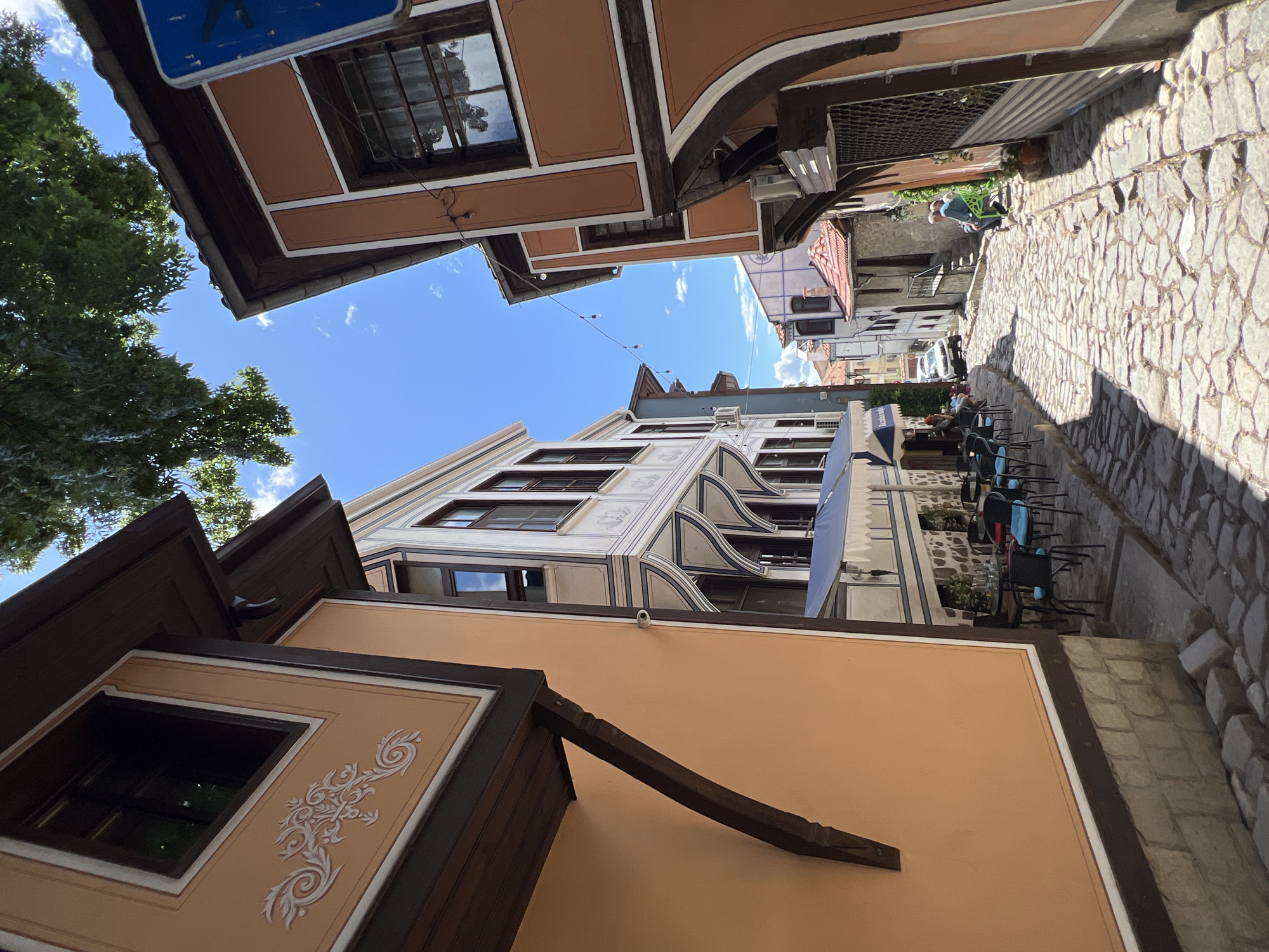 The image depicts a narrow, cobblestone street in Plovdiv, Bulgaria, flanked by traditional Bulgarian Revival-style buildings. The buildings feature distinctive architectural elements like overhanging eaves, painted facades, and decorative window frames. Several tables and chairs, suggesting a cafe or restaurant, are set up along the street. The sky is blue with scattered clouds, and sunlight casts shadows on the cobblestones. A few people are visible further down the street.