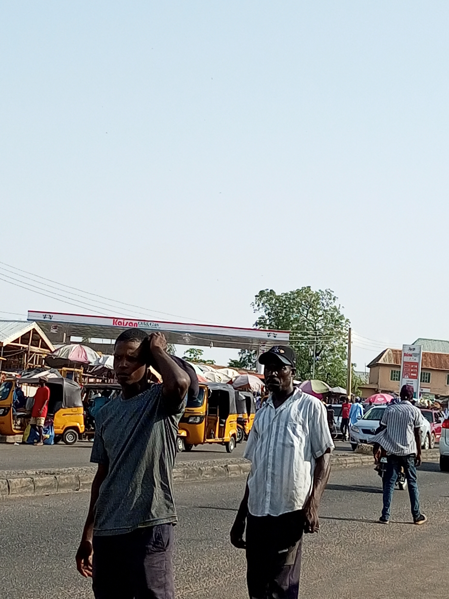The image shows a street scene in an urban area of Nigeria, likely a market or transportation hub. Two men are walking in the foreground. The man on the left is wearing a dark grey t-shirt and dark pants, with his right hand touching the back of his head. The man on the right is wearing a light-colored, vertically striped short-sleeved shirt and dark pants, with a black baseball cap. Behind them, several yellow auto-rickshaws, commonly known as "keke napep" or "Marwa", are parked or moving. A gas station with a prominent "Kaison Oil & Gas" sign is visible in the mid-ground, along with market stalls and umbrellas. Further in the background, buildings and more vehicles, including a white car and a red car, are present. The sky is clear and pale blue. The scene appears to be during daylight hours.