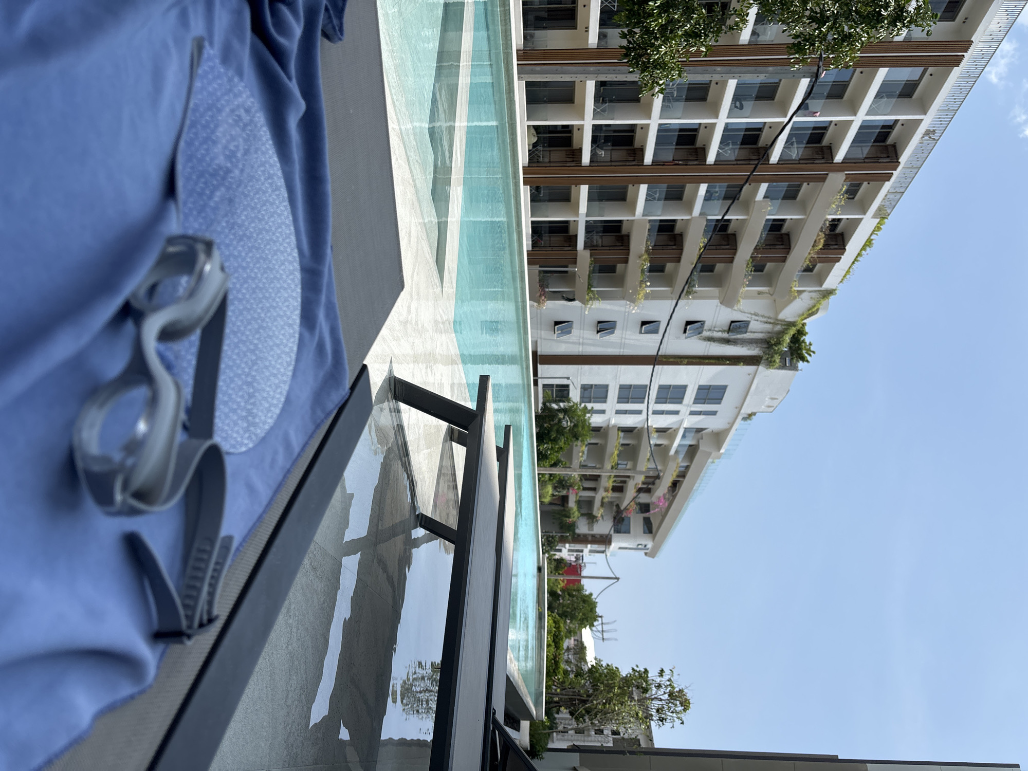 The image shows a scene from Choeng Thale, Thailand. The photograph is taken at an elevated angle, possibly from a lounge chair beside a pool. In the foreground, there is a pair of swimming goggles on top of a blue towel. The pool is next to the chair and extends outwards. Beyond the pool, multiple apartment buildings are visible with white facades, numerous windows, and balconies adorned with plants. The sky above the buildings is clear and blue. The focus of the photograph seems to be the building in the background.