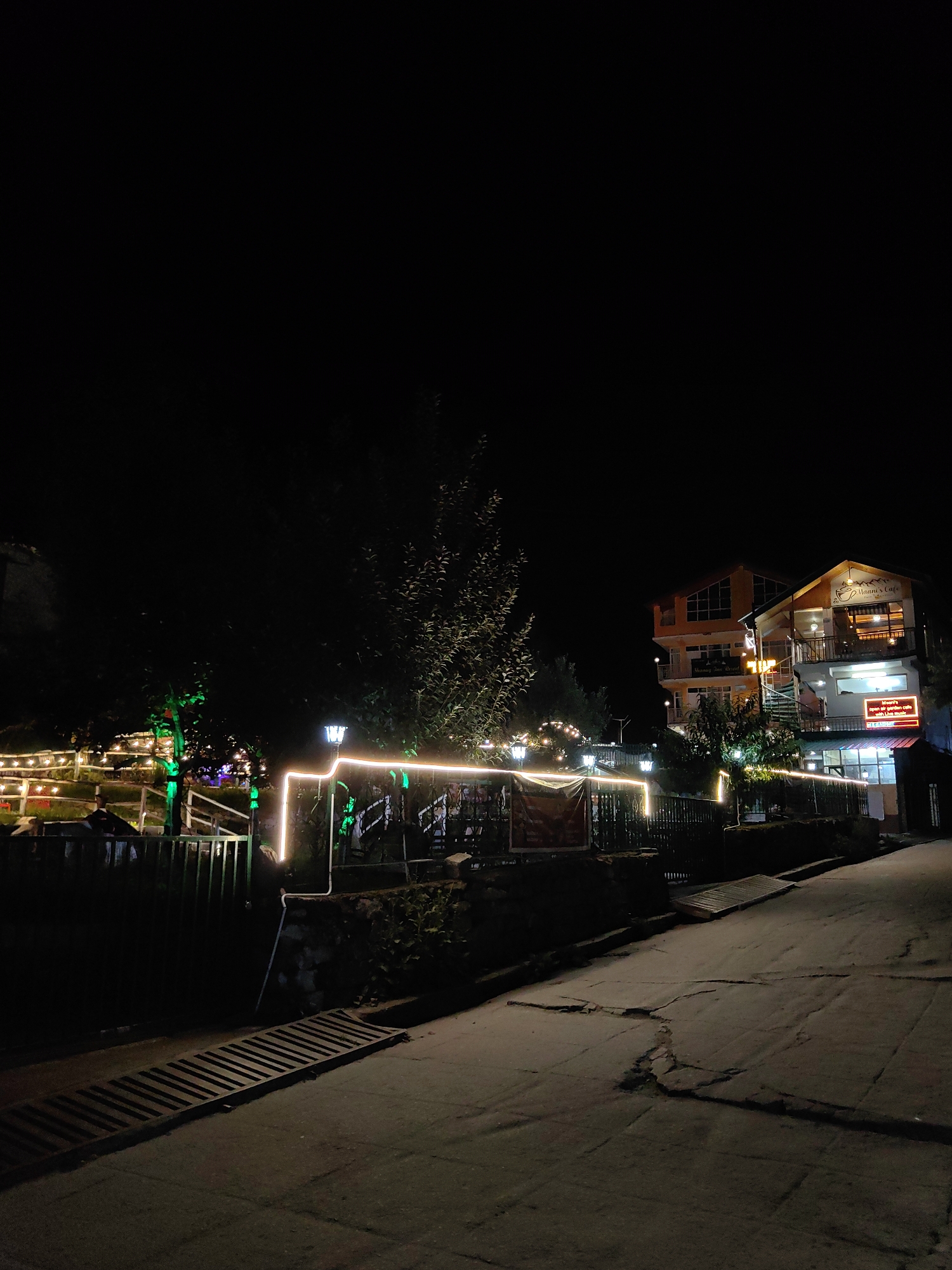 A night-time photograph captures a street scene in Barheta, India, illuminated by decorative lighting. In the background, two multi-story buildings are visible, one with an orange facade and the other with a white facade. A sign on the white building reads "Vaani's Cafe". The buildings are adorned with strings of lights, and a spiral staircase is visible on the side of the white building. A street lamp with a blue glow stands near the buildings. In the foreground, a road with cracks is present, and a metal grate lies on the side of the road. A low wall separates the road from an area with trees and more decorative lights, including green and white strands. Some patrons appear to be seated at tables in the dimly lit area with lights. The overall scene suggests a restaurant or cafe area open for business at night.