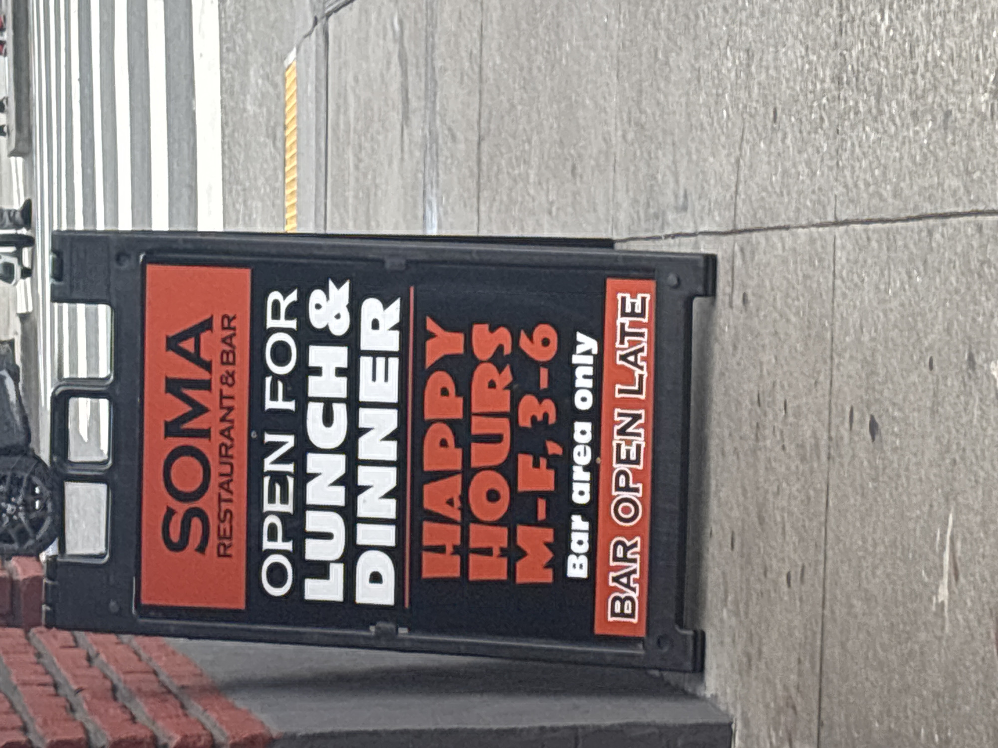 An A-frame sign advertising "SOMA RESTAURANT & BAR" is positioned on a sidewalk in front of a brick building. The sign is primarily black with red and white text. It states "OPEN FOR LUNCH & DINNER" in large white lettering. Below this, in large red lettering, it reads "HAPPY HOURS M-F, 3-6". Further down, in smaller white text, it says "Bar area only". The bottom of the sign has a red section with white text that reads "BAR OPEN LATE". The sidewalk is made of concrete with visible seams. In the background, a crosswalk with white stripes and a yellow tactile paving strip is visible, along with blurred figures of pedestrians and a portion of a vehicle. The overall scene suggests an urban commercial environment.