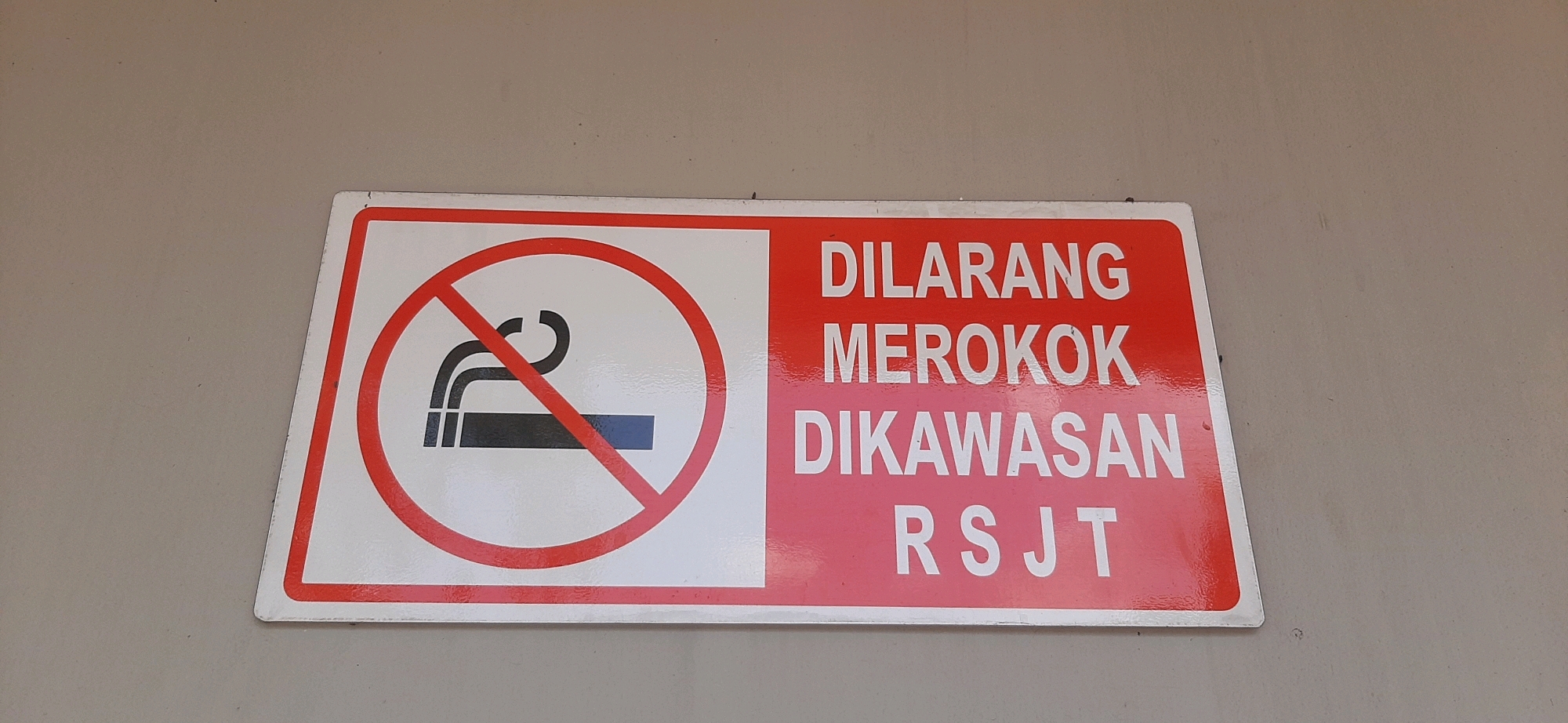 A rectangular sign with a white background on the left and a red background on the right is mounted on a beige wall. The left side of the sign features a no smoking symbol in red, with a stylized cigarette and smoke enclosed in a red circle with a red diagonal line through it. The right side of the sign has text printed in white capital letters. The text reads "DILARANG MEROKOK DIKAWASAN RSJT". This sign is located in the city of Tasikmalaya, Indonesia, likely at a hospital or healthcare facility given the abbreviation "RSJT" which often stands for "Rumah Sakit" (Hospital).