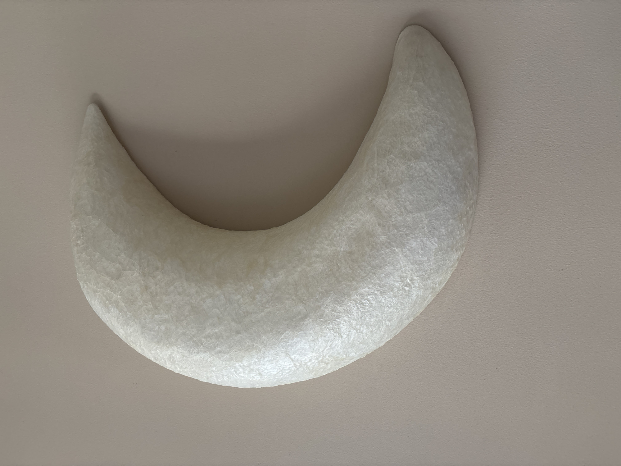 A textured, crescent-shaped object, resembling a moon, is affixed to a plain, light-colored wall. The object has a slightly iridescent, white surface with a rough, almost crackled appearance. The wall is a neutral, matte beige. The lighting casts subtle shadows that highlight the texture of the crescent. No people or other objects are visible in the scene. The location is not discernible from the image.