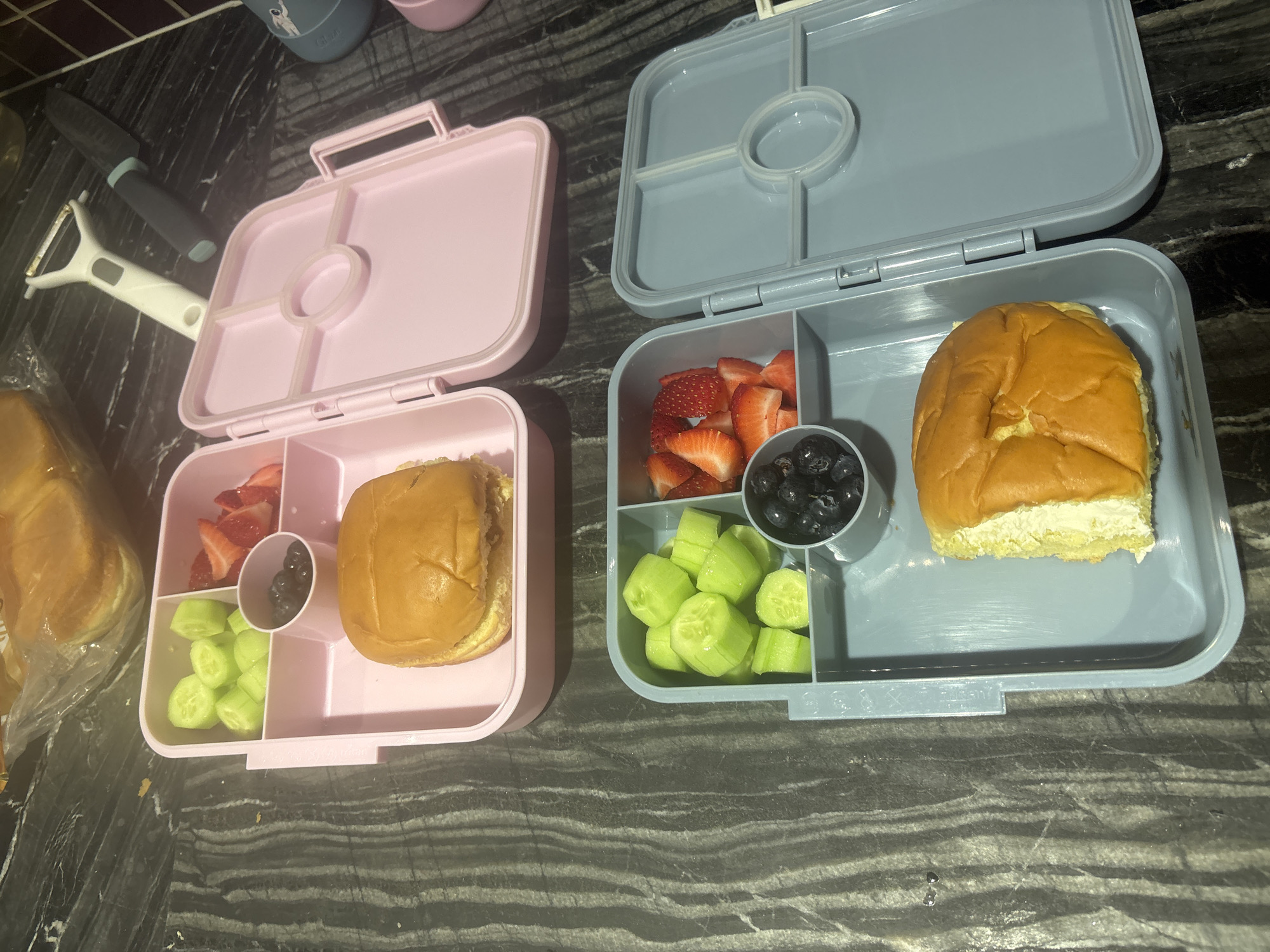 The image shows two open bento boxes filled with food. One bento box is pink and contains sliced cucumbers, sliced strawberries, blueberries, and a sandwich. The other bento box is blue-grey and contains sliced cucumbers, sliced strawberries, blueberries, and a sandwich. Both bento boxes are placed on a dark, speckled countertop. In the background, there are also some buns in a plastic bag and a white peeler and a black knife. The scene appears to be in a domestic kitchen setting, possibly in Dubai, United Arab Emirates.