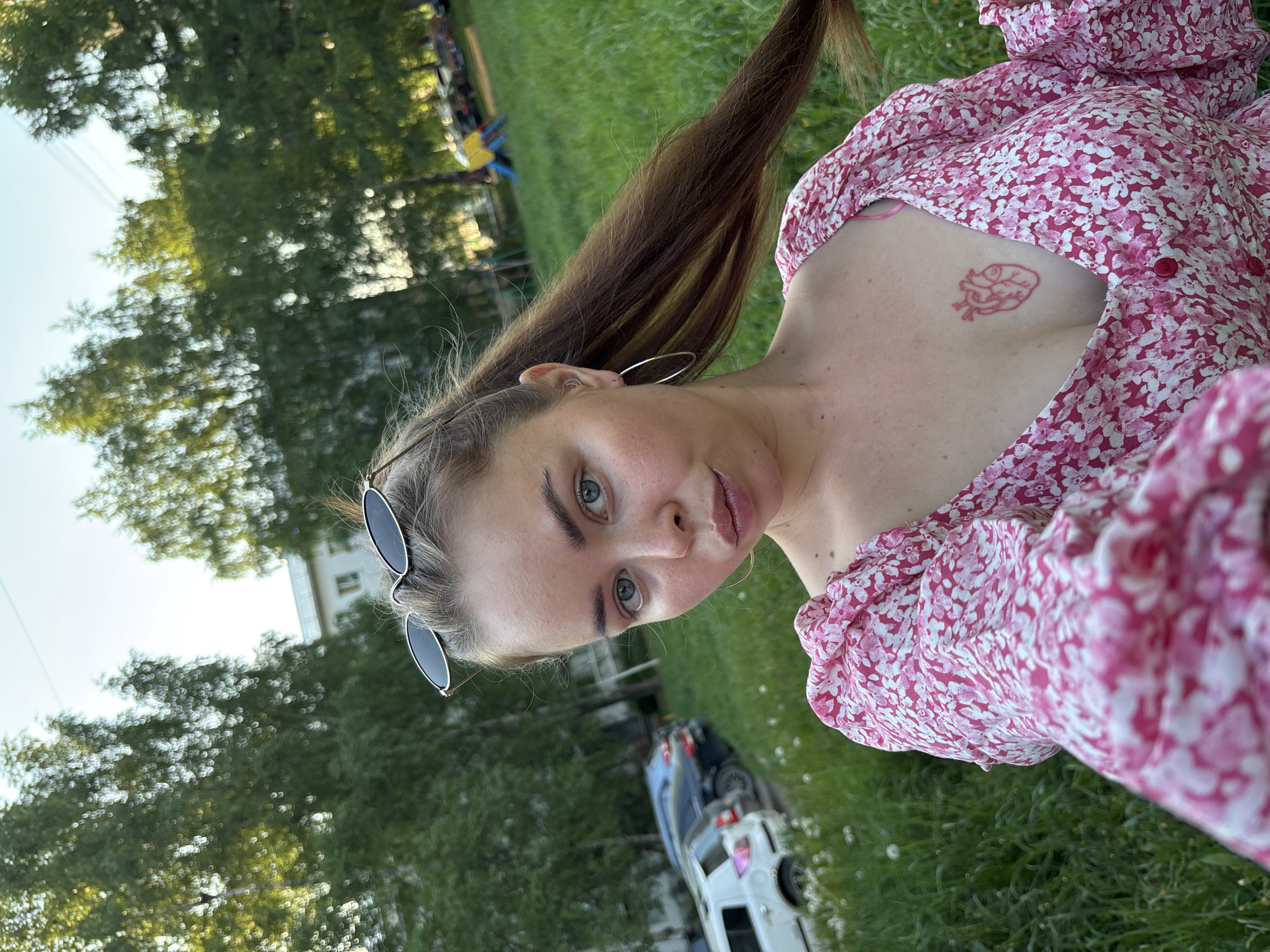A young woman with long brown hair, green eyes, and sunglasses perched on her head is shown in a rotated selfie outdoors. She is wearing a pink floral-patterned blouse. A red tattoo is visible on her chest. She wears hoop earrings. A car is visible parked nearby. The background appears to be a grassy area with trees. The location is Izhevsk, Russia.