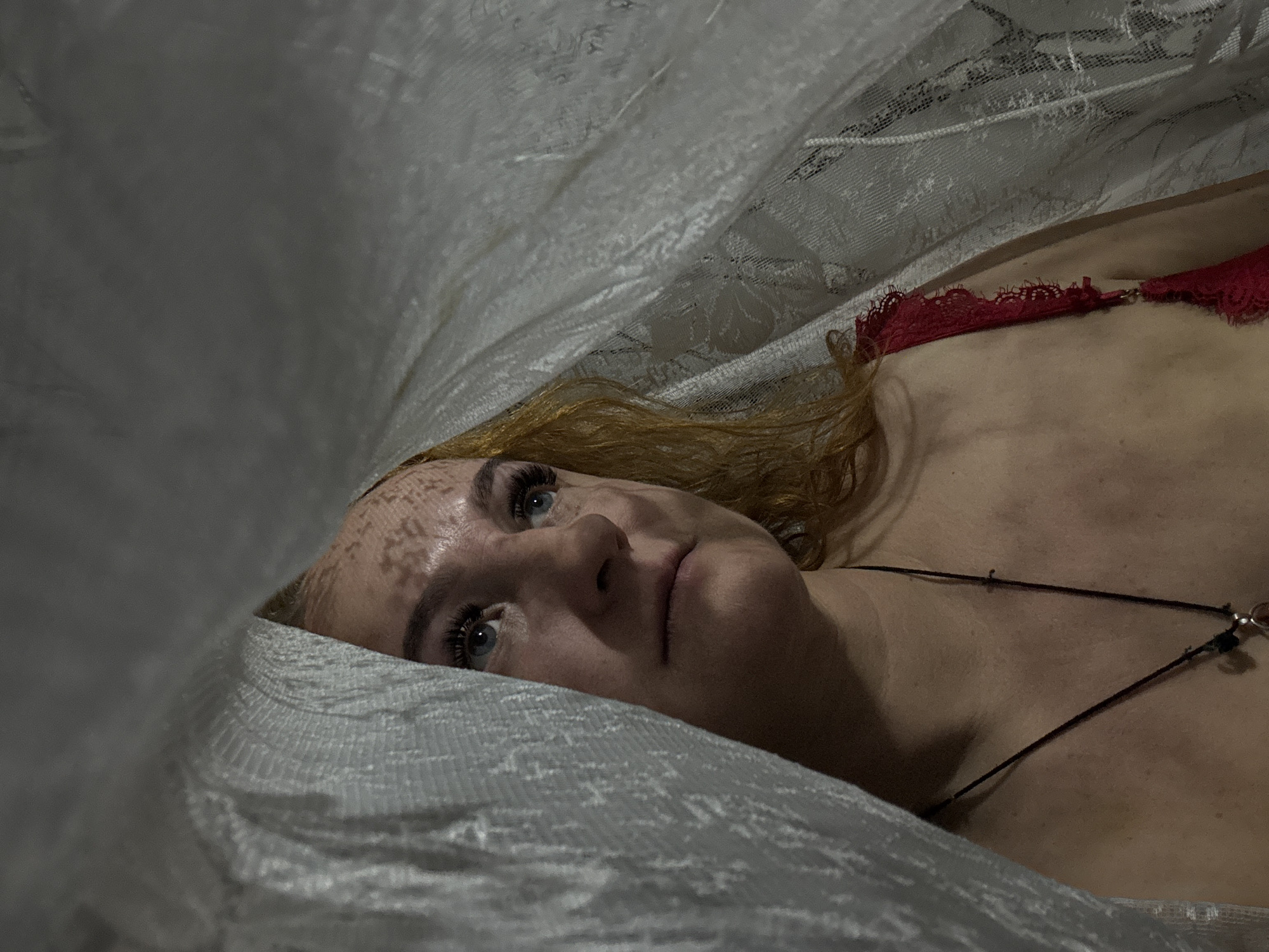 A fair-skinned, middle-aged woman with light red/blonde hair and blue eyes lies beneath a textured, semi-transparent white fabric. She wears a red, lacy garment on her upper body. A thin, black necklace adorns her chest. The lighting is dim and dappled, creating patterned shadows on her face. The background is obscured by the fabric. There is no verifiable indication of this image being taken in Chaikovsky, Russia.