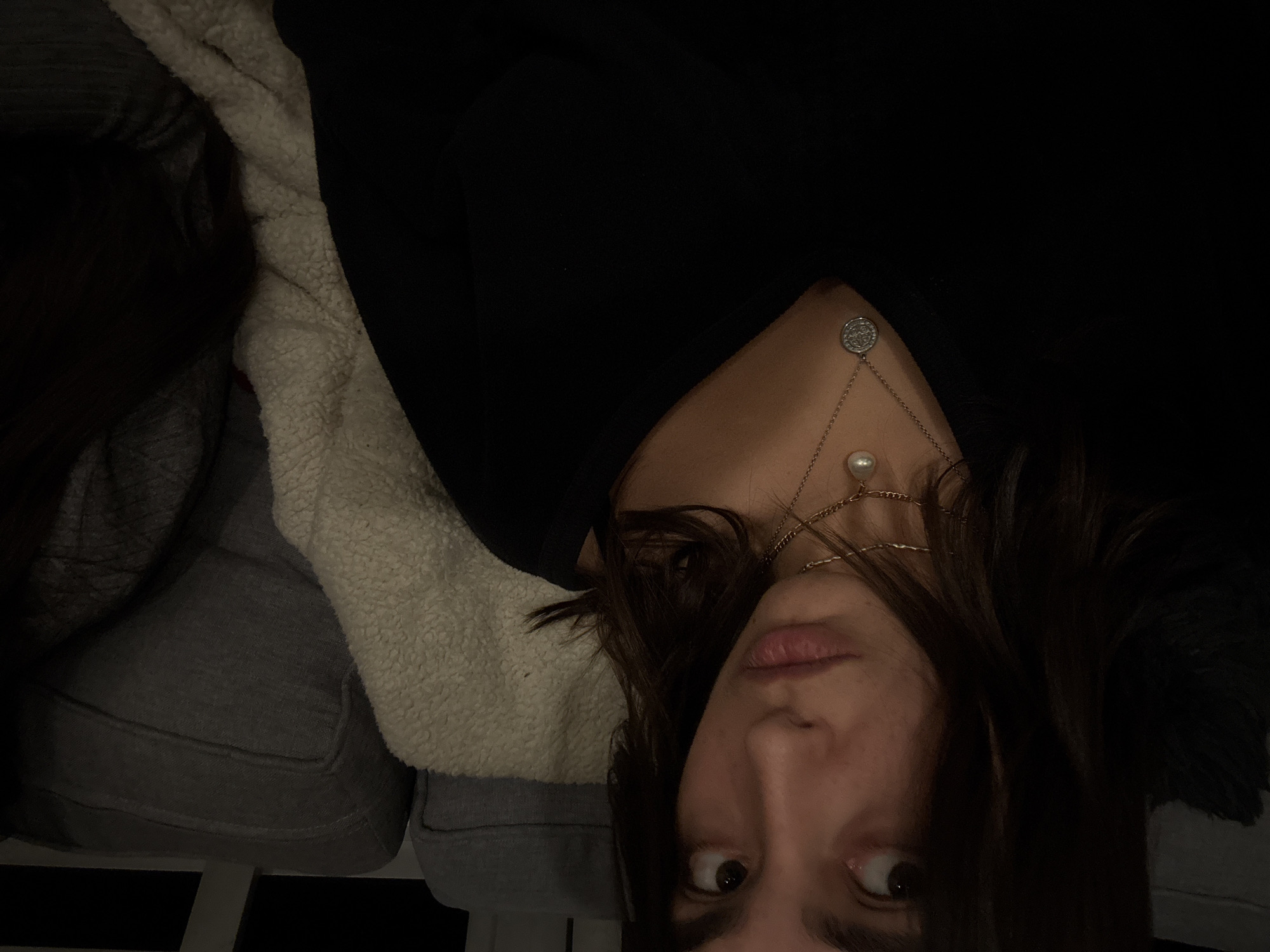 The image captures a close-up, sideways view of a person, likely a young woman, looking directly at the camera. She has dark hair that falls around her face and shoulders. She is wearing a black top or sweater and multiple layered necklaces. One necklace features a silver, coin-like pendant with an intricate, unreadable design, while another holds a round white pearl. A third, thinner gold chain is also visible.
The person appears to be resting on or next to a light-colored, fluffy, possibly sherpa-lined blanket or cushion, with a dark grey upholstered surface, likely a sofa or chair, visible in the background. The lighting is dim, suggesting it is evening or night. Her expression is neutral, with her eyes wide and gazing directly at the viewer. The overall mood is casual and intimate, possibly a selfie or a relaxed portrait taken in an indoor setting. The location context provided is Mexico City, Mexico.