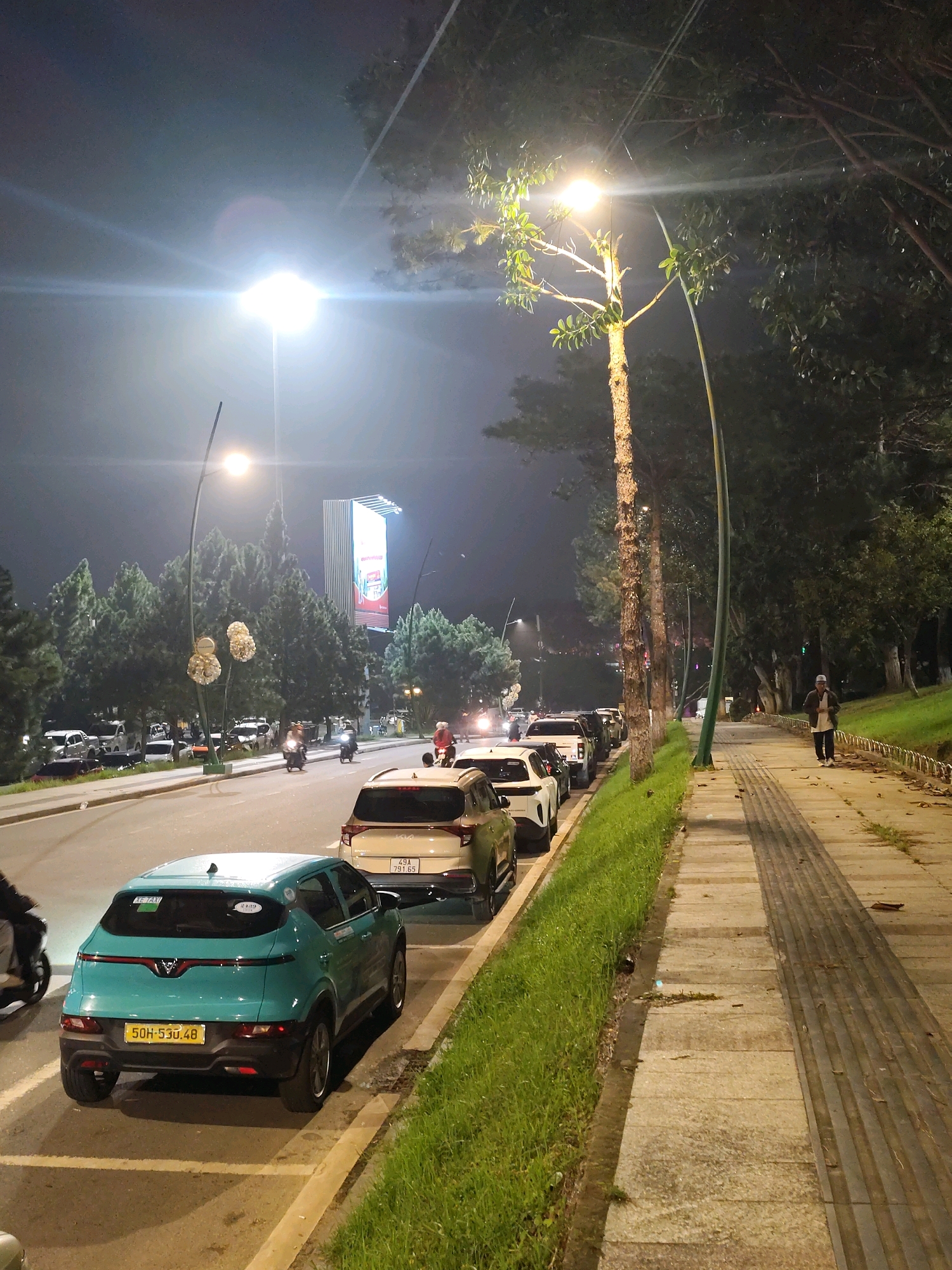 The image depicts a nighttime scene on a street in Da Lat, Vietnam. A turquoise car is parked on the right side of the road, facing away from the viewer. A person is walking on the sidewalk on the right side of the image, and several other cars are parked along the street. Trees line the sidewalk, and streetlights illuminate the scene. In the background, a large billboard is visible. The overall atmosphere is that of a quiet urban night.