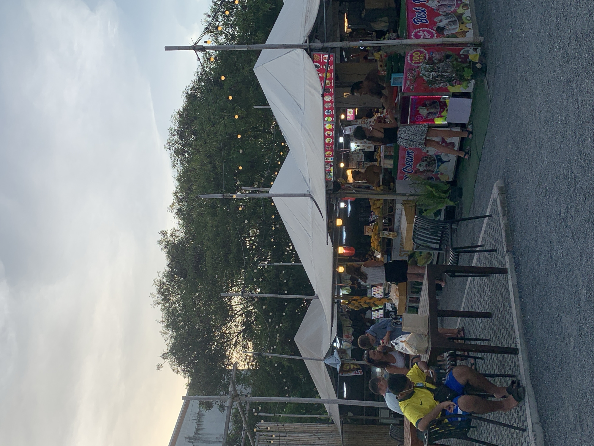 The image depicts an outdoor market scene in Ko Samui District, Thailand, during the late afternoon or early evening. The sky is overcast with a soft, diffused light. A row of white tents with peaked roofs is set up, housing various market stalls. One stall prominently displays colorful images of ice cream and has text that reads "Ice Cream" and "Best". Several people are visible. In the foreground, a young man wearing a yellow t-shirt and blue shorts sits at a dark wooden table, looking at his phone. Other diners are seated at the same table and another table to the left. In the mid-ground, a woman in a white top and black skirt stands at an ice cream stall, interacting with a vendor. Another stall to the left appears to be selling fruit, with bunches of yellow fruit hanging from it. The ground is covered in small grey gravel. Strings of yellow lights are strung between poles and over the tents, adding to the market atmosphere. Lush green trees form a backdrop behind the market stalls.