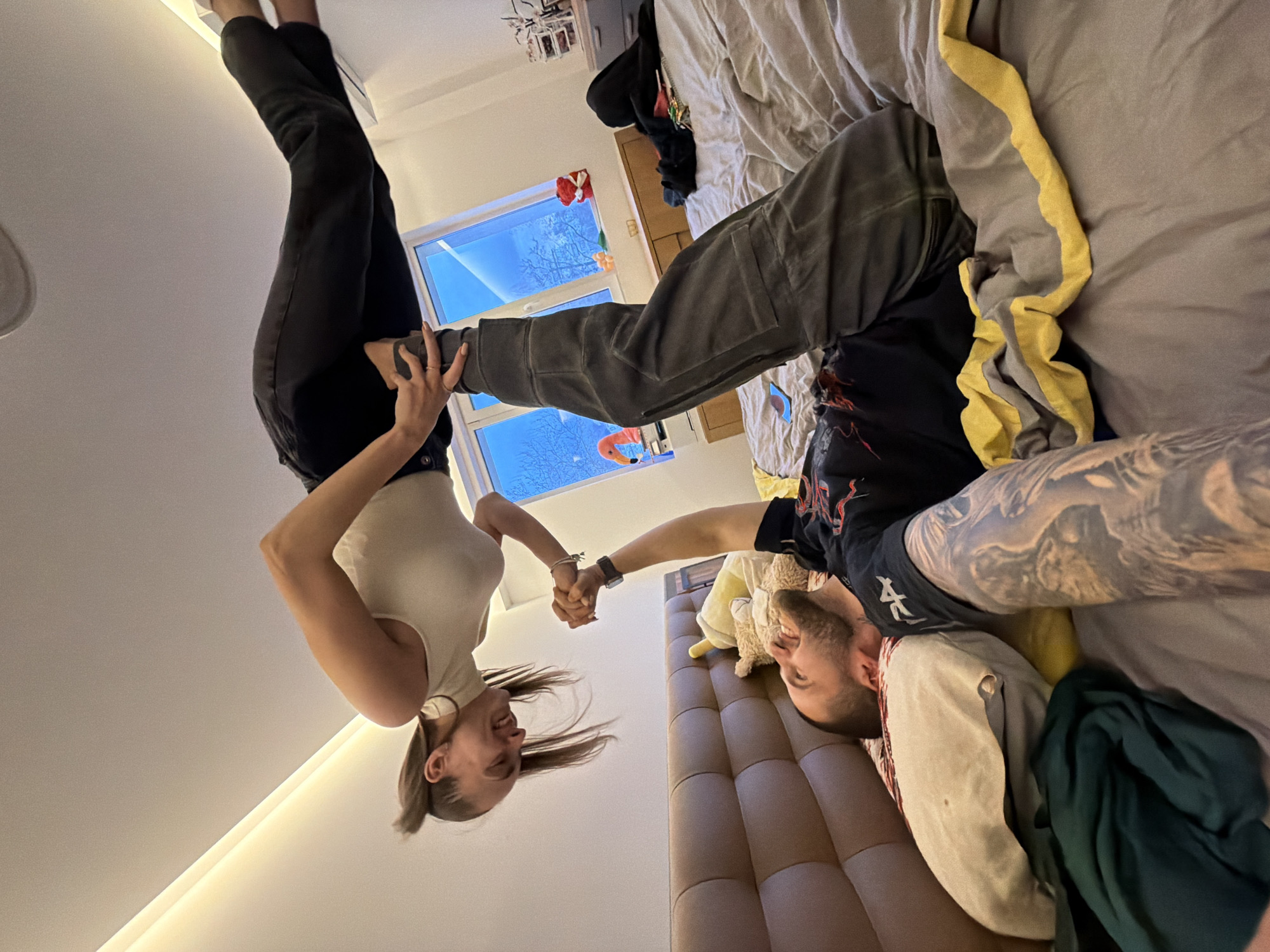 Two people are oriented upside down in the image. A woman wearing a white tank top and dark pants is hanging upside down, supporting herself with her hands clasped with a man lying on a bed. The man has tattoos on his arms and is wearing a dark T-shirt and light-colored pants. The background features a window showing a cityscape with bare trees. The scene appears to be indoors, in a bedroom with a bed, pillows, and other personal items. It is impossible to determine if the image was taken in Sofia, Bulgaria without further information.