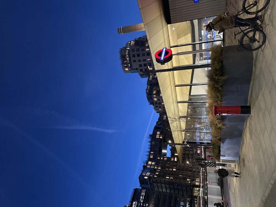 The image depicts the area around Battersea Power Station Underground station in London, United Kingdom, at twilight or early evening.
The scene features a modern London Underground station entrance on the right, identifiable by its illuminated red and blue "UNDERGROUND" roundel sign. The station building has a light-colored, angular roof and extensive glass paneling. To the left of the station entrance, a large rectangular planter filled with dry, wispy vegetation separates the building from the paved walkway. A traditional red British pillar box stands centrally in the foreground, adjacent to this planter.
On the right, a man with a beard, wearing a brown jacket, dark trousers, and a white hat or beanie, stands beside a black bicycle, holding a mobile phone. Further left, another individual in a dark jacket and light trousers walks away from the viewer on the paved area. A third person on a bicycle is partially visible in motion on the far left.
In the background, a series of modern, multi-story buildings, predominantly residential with numerous illuminated windows, create a developed urban skyline. A tall, light-colored industrial chimney, characteristic of the Battersea Power Station, is distinctly visible on the far right horizon, rising above the station building. The sky is a deep blue, showing two prominent white contrails from aircraft. The ground is paved throughout the visible area.