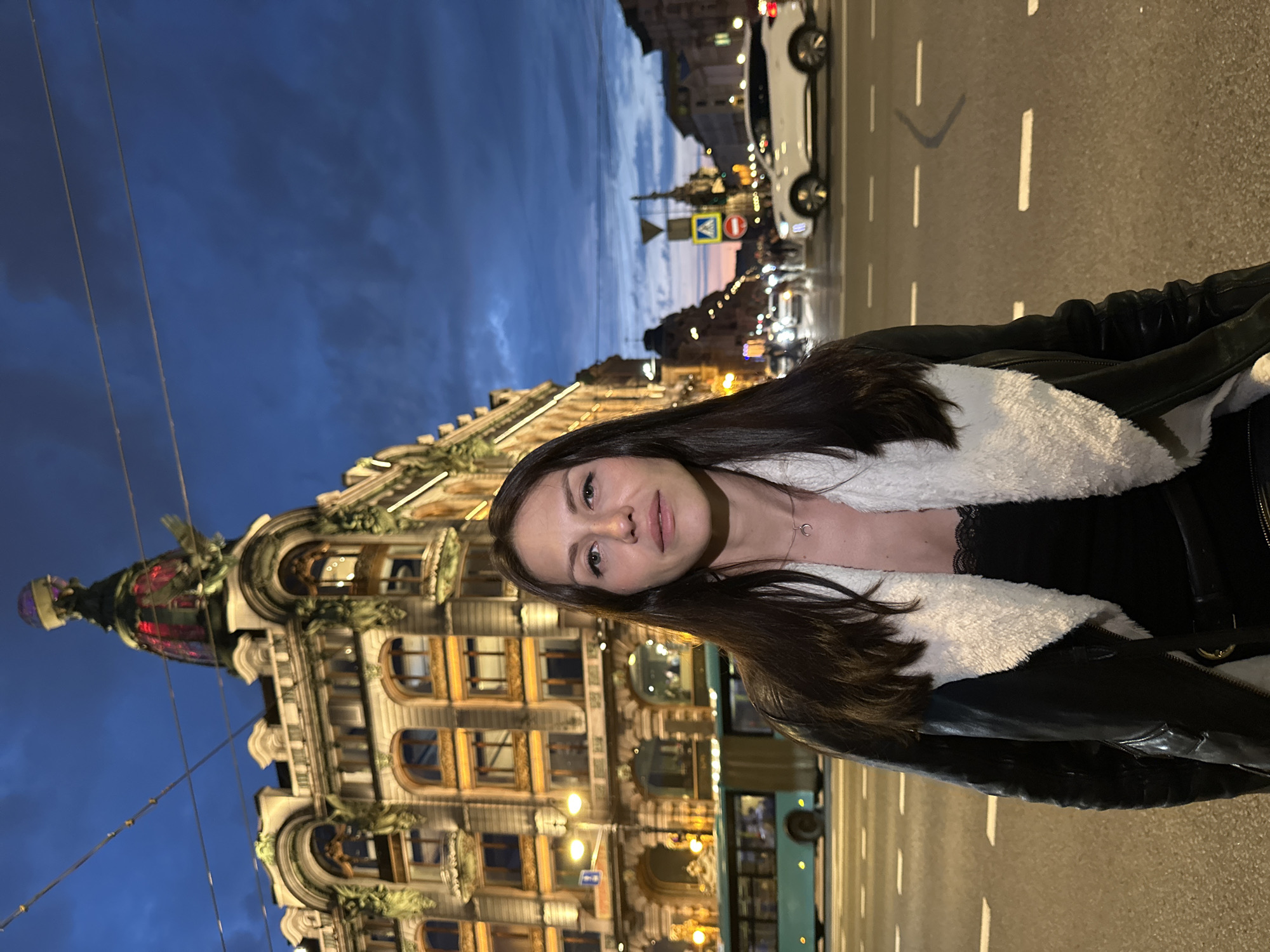 A woman stands in the middle of a street in Saint Petersburg, Russia, at dusk. She is wearing a black leather jacket with a white fur collar over a black lace top, and a crescent moon pendant necklace. The street is lit by streetlights and the illuminated windows of a large, ornate building behind her. The building features a dome-shaped roof with a globe on top and decorative architectural elements. In the background, traffic, including a bus and cars, moves along the street. The sky is a deep blue with hints of orange and pink from the setting sun, and some clouds are visible. The woman is looking directly at the camera with a neutral expression.