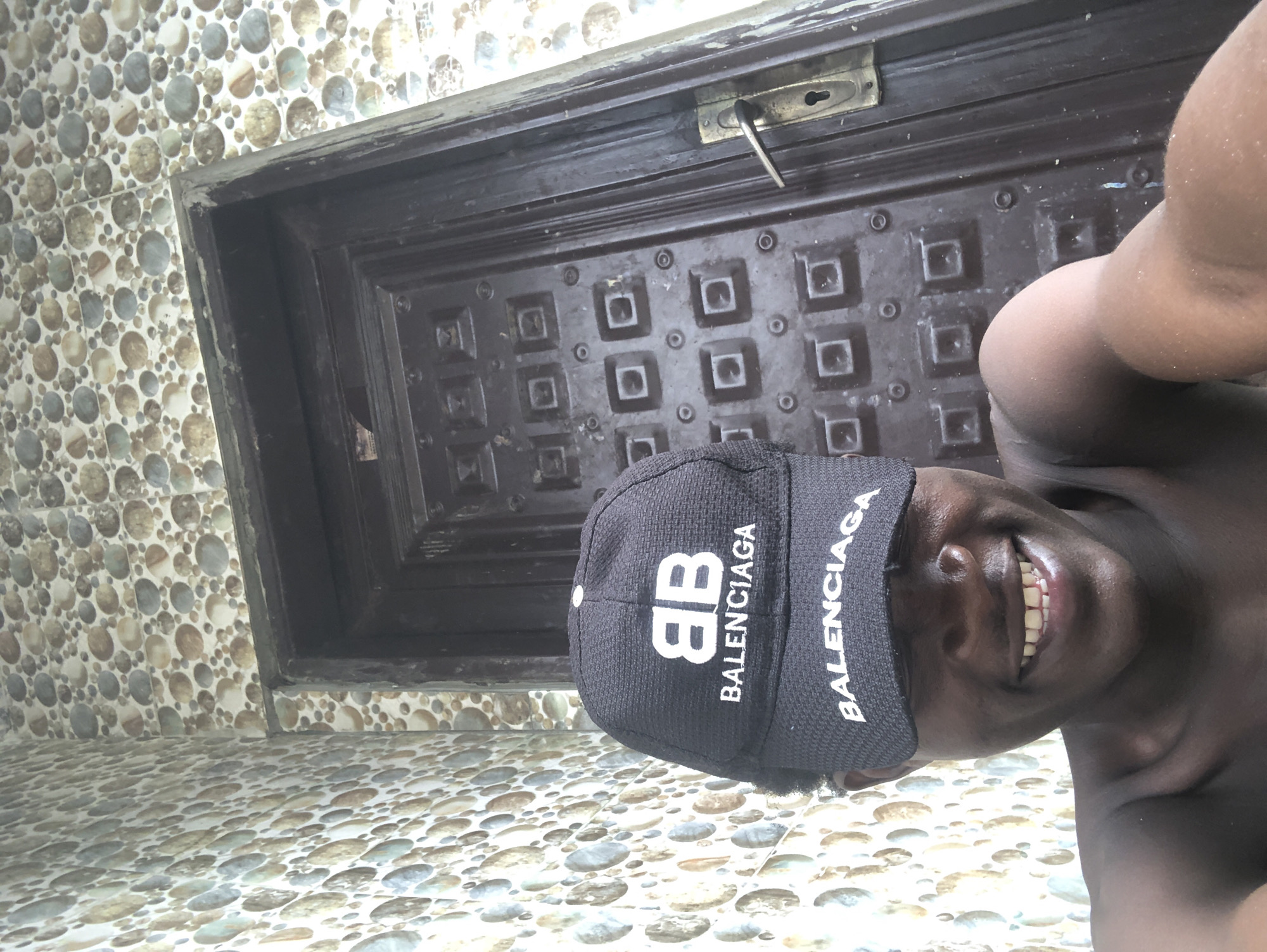 In Ado, Nigeria, a person is taking a selfie in front of a dark, intricately designed wooden door. The door has a metal handle and a keyhole, and is set within a wall covered in colorful, circular patterned tiles. The person is wearing a black Balenciaga cap, which is partially obscuring their face. They are smiling broadly, revealing their teeth. The setting appears to be an outdoor or semi-outdoor area, possibly a porch or entrance.