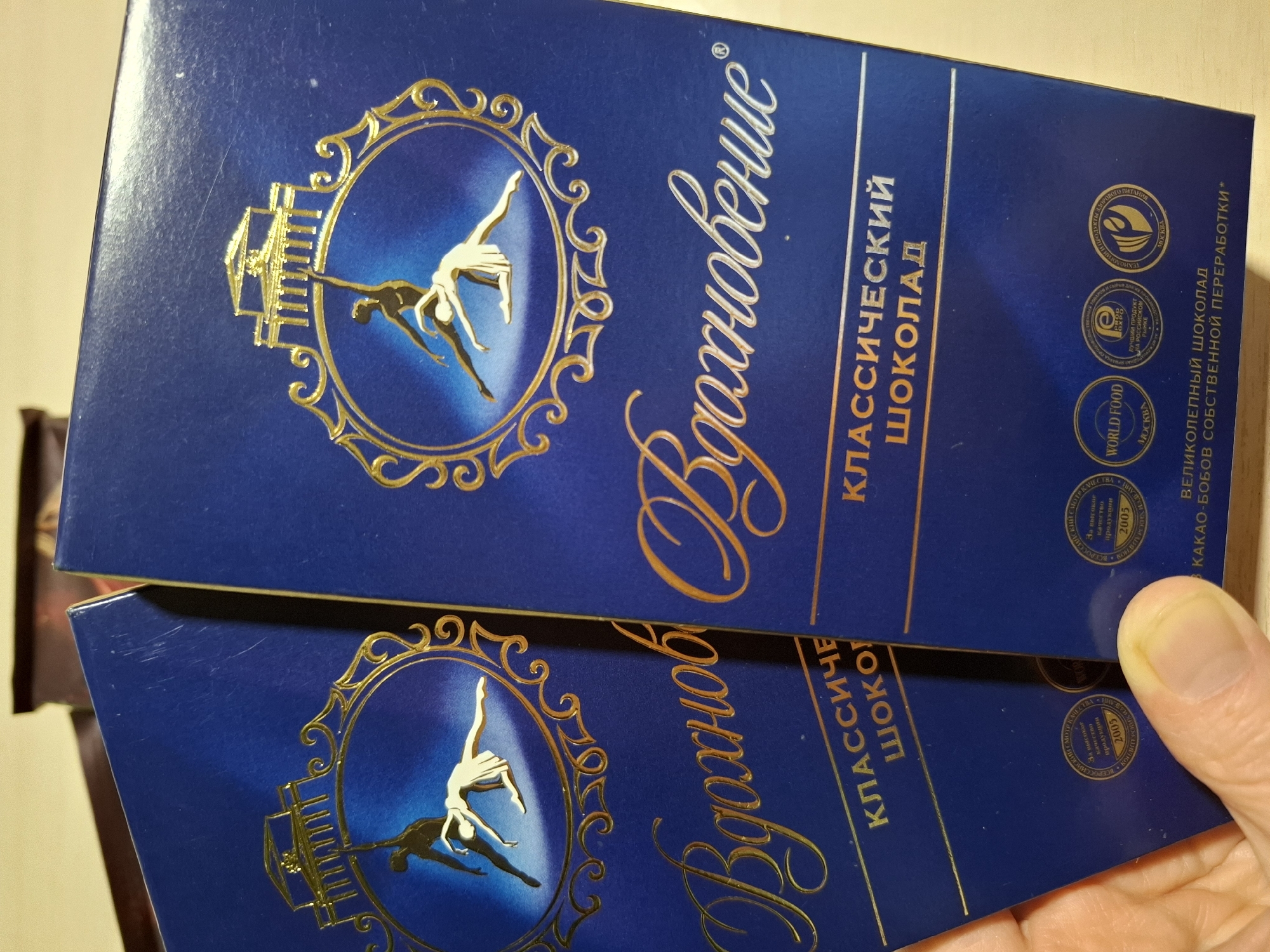 The image shows two blue chocolate bars from the brand "Vdokhnovenie" (Inspiration). The bars are positioned diagonally, with the left bar slightly in front of the right. Both bars have a golden emblem at the top featuring two figures in a ballet pose, set within an ornate frame. Below the emblem, the word "Vdokhnovenie" is written in large, elegant golden script. Further down, the text "КЛАССИЧЕСКИЙ ШОКОЛАД" (Classic Chocolate) is visible in golden lettering. The lower part of the bars displays various circular stamps and text, some of which are partially obscured. One stamp on the left bar includes the text "2005" and what appears to be "WORLD FOOD". The image is taken from a slightly elevated angle, with a person's thumb and finger holding the left chocolate bar. The background is blurred but appears to be a light-colored surface. There is no indication of the city of Tyumen, Russia in the image.