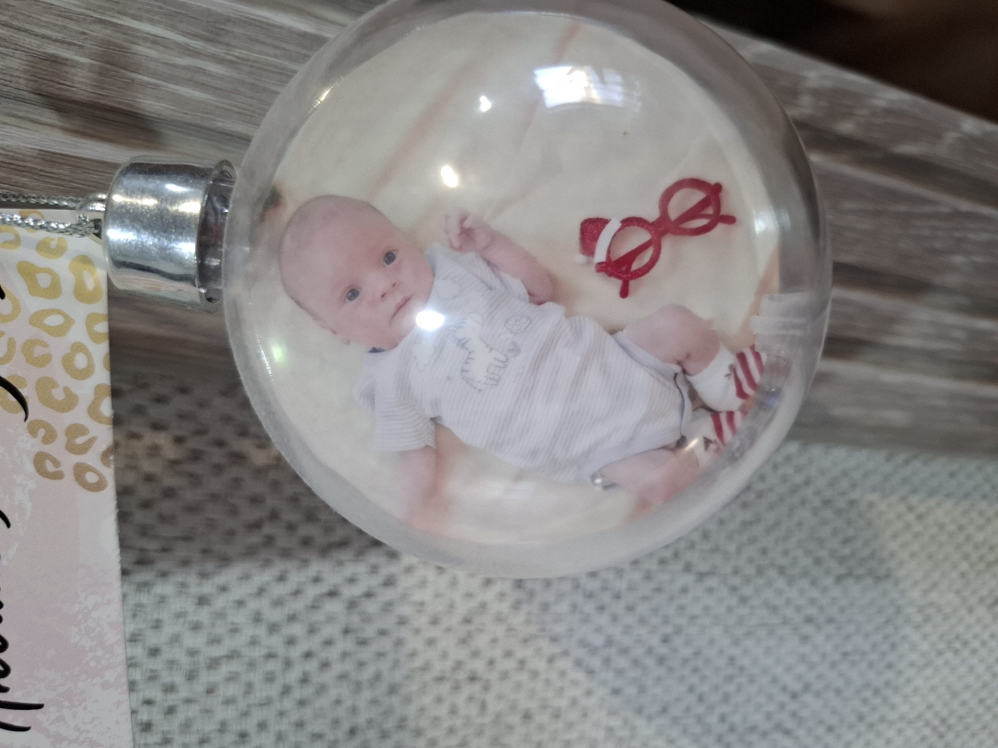 A clear Christmas ornament containing a photograph of a baby lying on its back, wearing a striped onesie and socks. Beside the baby in the photo are small red glasses. The ornament is positioned on a textured surface, with a metallic top. A printed tag is partially visible on the left side. The location of Амзя, Russia, is not relevant.