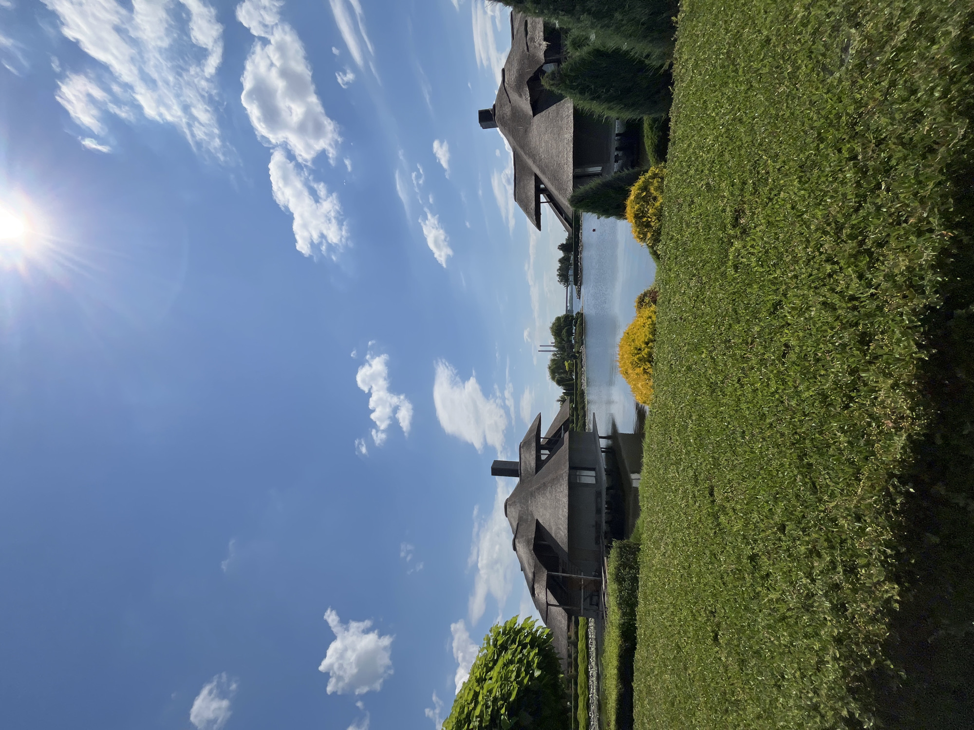 The image depicts a serene waterside landscape under a bright, sunny sky with scattered white clouds. In the foreground, meticulously manicured green hedges and bushes form a lush lawn. Beyond the lawn, a tranquil body of water reflects the sky and surrounding structures. Two distinctive buildings with thatched roofs and dark walls stand on stilts over the water, their architectural style reminiscent of thatched bungalows. One building is situated to the left, partially obscured by a large, leafy green tree. The other building is positioned to the right, with several cone-shaped evergreen trees flanking it. In the distance, across the water, industrial structures and a bridge are visible, suggesting the presence of a city. The overall scene evokes a sense of peaceful retreat, potentially a resort or a private residence, located near an urban area. This location is identified as the Dnipro River embankment in Dnipro, Ukraine. There are no people visible in the image.