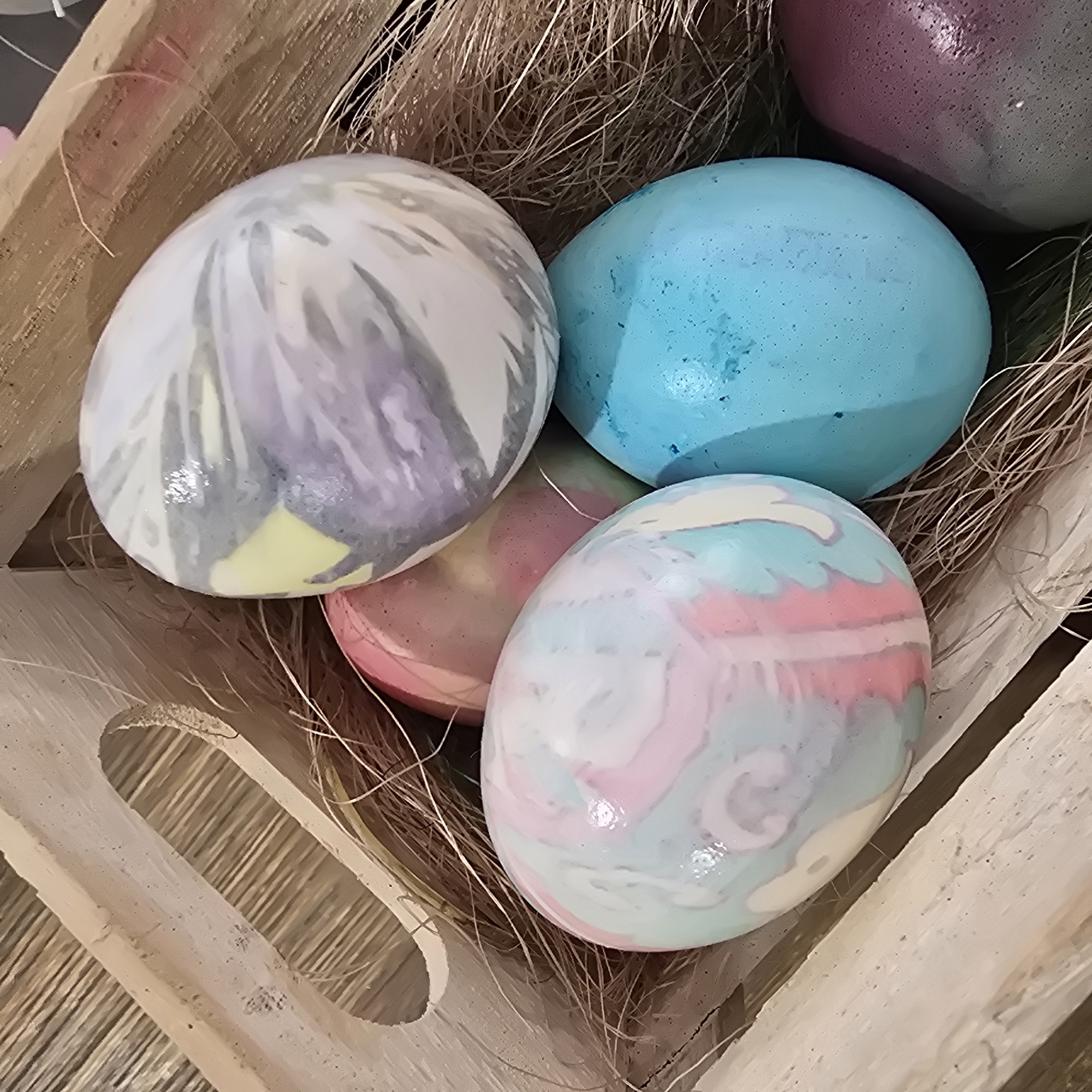 A close-up shot depicts a collection of decorated eggs in a wooden crate. The eggs are painted in various colors and patterns, including marbled designs and solid colors. The crate appears to be filled with straw or similar nesting material. The image lacks context that would connect it to Plovdiv, Bulgaria. No people, actions, or interactions are visible.