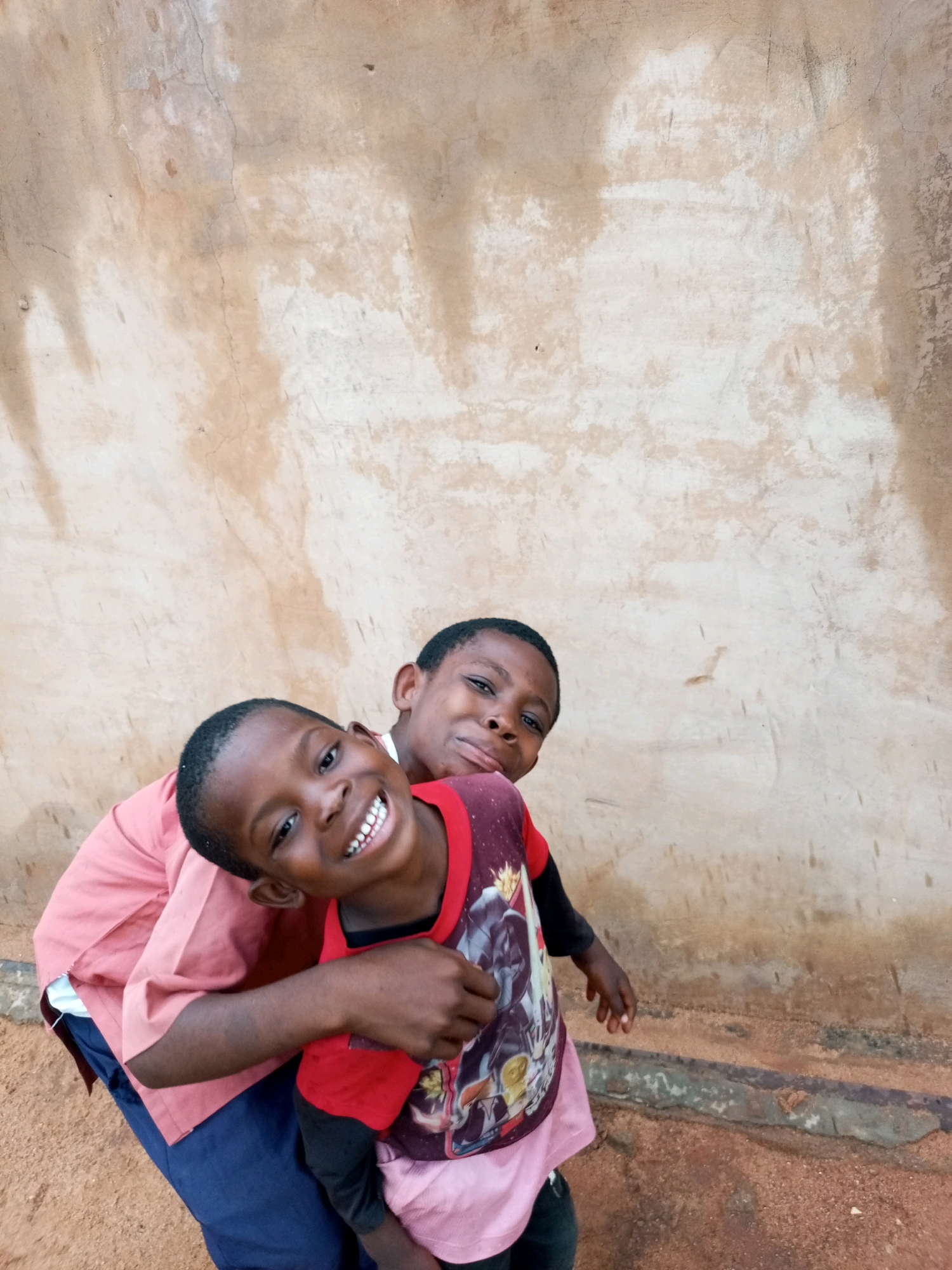 Two young Black boys are posing for a photo against a textured, light brown wall with a roughly plastered surface. The boy in the foreground is smiling widely, showing his teeth, and leaning towards the camera. He is wearing a red t-shirt with a graphic print and black sleeves, and a pink undershirt. The other boy is behind him, with his head tilted back and a playful expression. He is wearing a pink collared shirt over a white undershirt. They are standing on a dirt or sandy ground. The image was captured in Tempure, Nigeria.