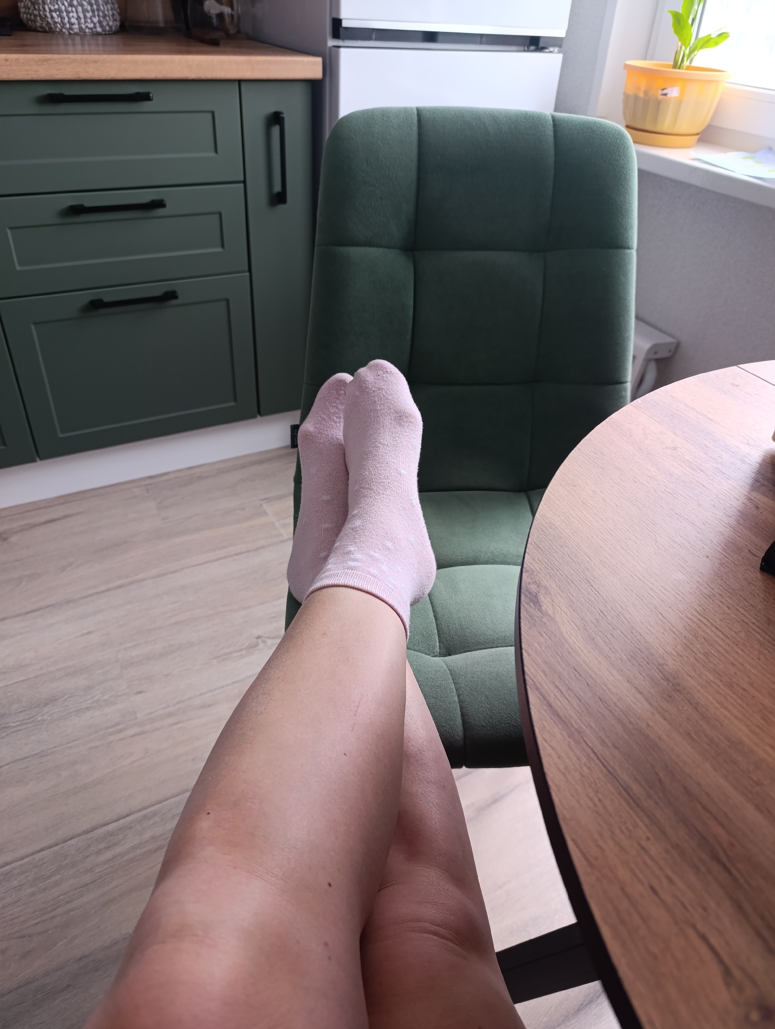 The image shows a pair of bare legs crossed and resting on a green upholstered chair. The feet are clad in pale pink socks with a small dotted pattern. A round wooden table is partially visible to the right of the chair. In the background, there are green kitchen cabinets with black handles and a white refrigerator. A windowsill with a potted plant is visible in the upper right corner. The scene appears to be set indoors, possibly in a kitchen or dining area.
