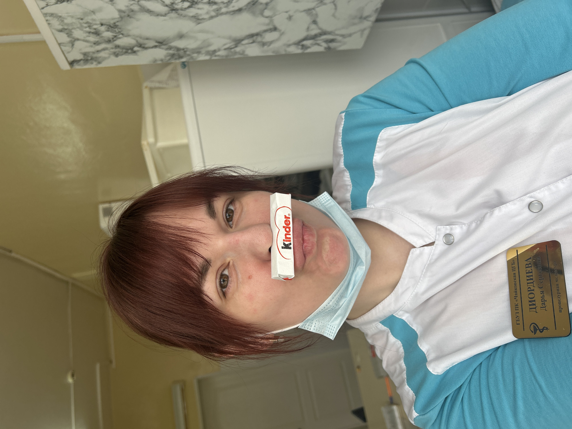 A woman with reddish-brown hair has a Kinder chocolate bar balanced on her nose. She is wearing a light blue medical mask pulled down under her chin and a white lab coat with light blue trim. Her name tag reads "ДИОРДИЕВА Дарья Сергеевна." The text on the badge identifies it as belonging to the ГБУЗ ПК "Чайковская ЦГБ" (State Budgetary Healthcare Institution of the Perm Territory "Tchaikovsky Central City Hospital"), indicating the city of Фоки, Russia. The background appears to be inside a medical or office setting. There's a marble-patterned surface and part of a white appliance (refrigerator) visible.