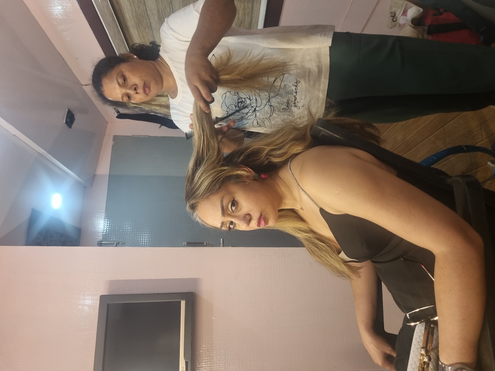 An indoor scene captures two women engaged in a hairdressing activity. In the foreground, on the right, a woman with long, highlighted blonde and light brown hair is seated, looking towards the viewer with a neutral expression. She is wearing a black top or dress and a thin necklace, with a pink earring visible.
Behind and slightly to the left of her, another woman, presumably the hairdresser, stands, focused on the client's hair. She has dark hair, pulled back, and is wearing a white t-shirt with a stylized black rose graphic and text that appears to read "live yourself" (the word "yourself" is clearly visible). She is also wearing dark pants. Her hands hold a comb and scissors, actively working on the client's hair, which she is sectioning and trimming.
The setting appears to be a salon or a designated area for hair services. The walls are painted a pale pink or light mauve color, with a section of textured gray or blue paneling visible in the background. A bright light source is visible on the upper left wall, reflecting off a mirror or shiny surface. The floor is a light brown, possibly wood or laminate. In the bottom left corner, part of a dark-framed monitor or screen with an "LG" logo is visible. The overall lighting suggests an indoor, artificially lit environment. Both individuals appear calm and focused on the task at hand.