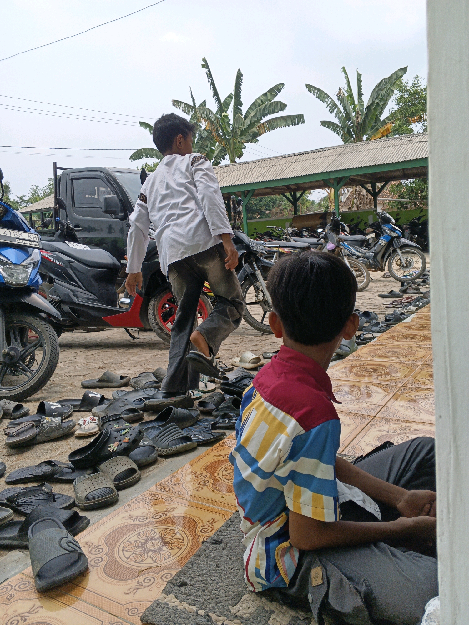 In Gedung Nyapah, Indonesia, a boy wearing a white shirt with decorative trim and grey pants walks away from the camera, stepping over numerous pairs of black and grey sandals scattered on a tiled courtyard. He is near a black utility truck and several parked motorcycles. In the foreground, another boy, facing away from the camera and wearing a colorful striped shirt with red, yellow, blue, and white stripes, sits on the ground. The environment includes a covered outdoor seating area with a corrugated metal roof, supported by green posts, and lush green banana trees in the background under a cloudy sky.