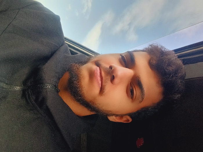 A portrait photograph, taken in Kafr Sousa Municipality, Syria, captures a young male from the chest up, seated inside a vehicle. The subject, likely in his late teens or early twenties, has medium brown skin, dark curly hair, a short beard, and dark eyes. He is wearing a black hooded jacket with a black zipper partially fastened, and looks directly at the camera with a neutral expression. To the subject's left, a window reveals a clear blue sky with sparse white clouds. To his right, the dark interior of the vehicle is visible, with a small, blurry red object positioned in the upper right corner. No other individuals, objects, or actions are discernible within the frame.