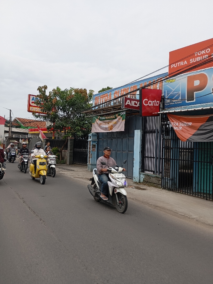 The image shows a street scene in Kecamatan Kutawaringin, Indonesia (latitude: -6.974, longitude: 107.542). Six people are riding motorbikes on a paved road, traveling from left to right. A white motorbike with a blue license plate is in the foreground, with a man wearing a blue shirt and a black cap riding it. The man is wearing a blue shirt and a black cap. The road is flanked by sidewalks and buildings. There are several shops with signs in the background. The shops have blue and white walls and black metal gates. There is a green tree in the left background. The sky is overcast and gray. The scene is in daytime. The image was likely taken on a street near a commercial district.