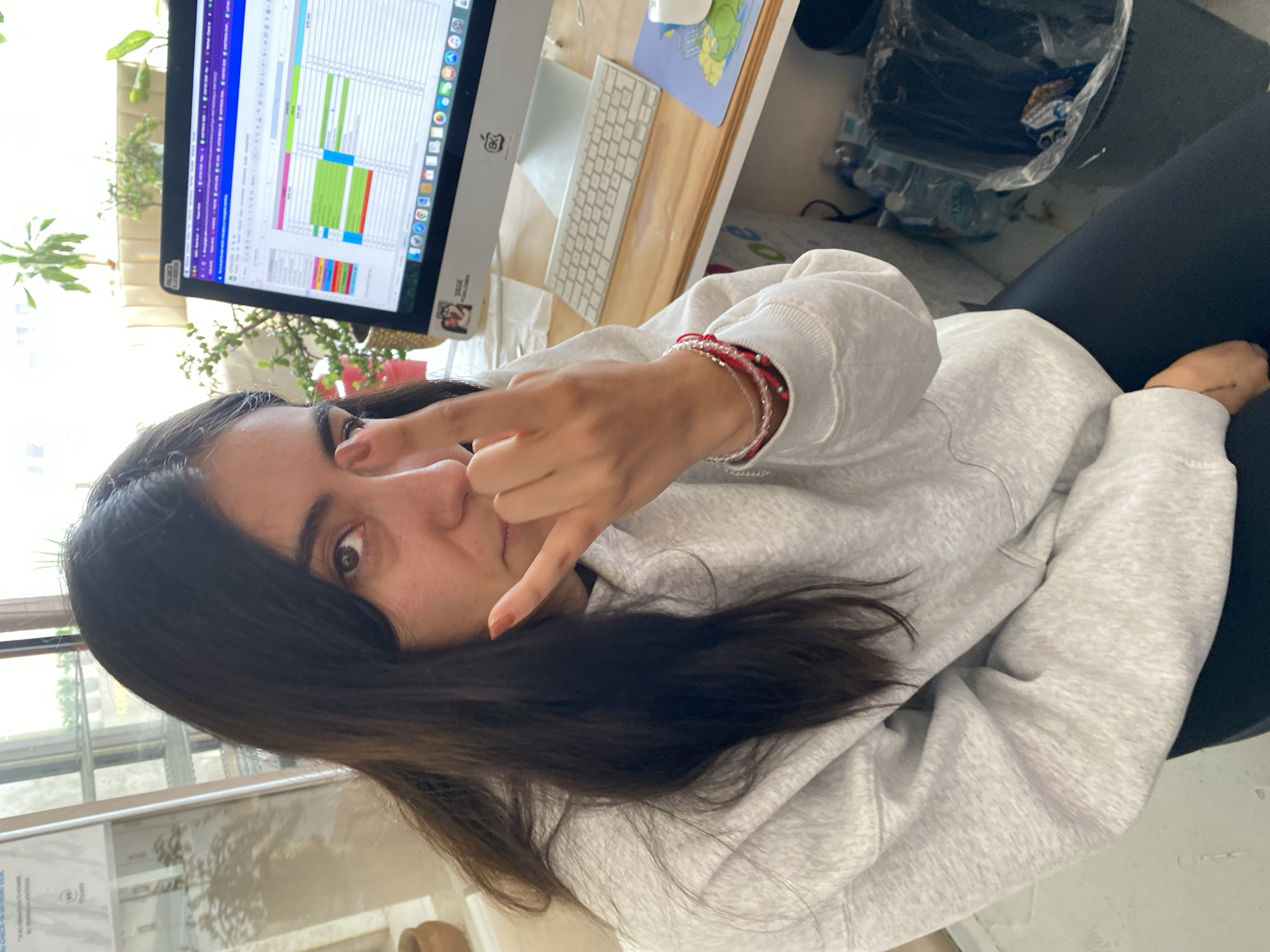 A young woman with long, dark hair is the central subject of this casual portrait, presented rotated 90 degrees clockwise. She wears a light grey hooded sweatshirt and is captured mid-gesture, holding her right hand up in a "rock on" sign (index and pinky fingers extended) in front of her face. Her right wrist is adorned with multiple red and pink string bracelets, and her fingernails are painted a reddish-brown. Her dark eyes look directly at the viewer with a neutral to slightly playful expression.
She appears to be seated at a wooden desk during the day, indicated by bright natural light streaming from a window visible behind a computer monitor. The monitor displays a detailed, colorful spreadsheet or scheduling application, along with various software icons. A white keyboard and a white computer mouse rest on the desk beside the monitor. Behind the screen, several green potted plants add greenery to the indoor setting.
Below and to her right, a black plastic bag and a plastic water bottle are visible on a lower surface or the floor, next to her legs, which are clad in dark leggings. A small, unidentifiable black tattoo is present on her left wrist. On the lower right frame of the computer monitor, a sticker clearly reads "SIGUE ADELANTE," which is Spanish for "Keep Going" or "Go Forward." Another blurry sticker is on the lower left of the monitor frame. The scene suggests a relaxed moment taken during what could be a work or study period in an office or home office environment in Puebla, Mexico.