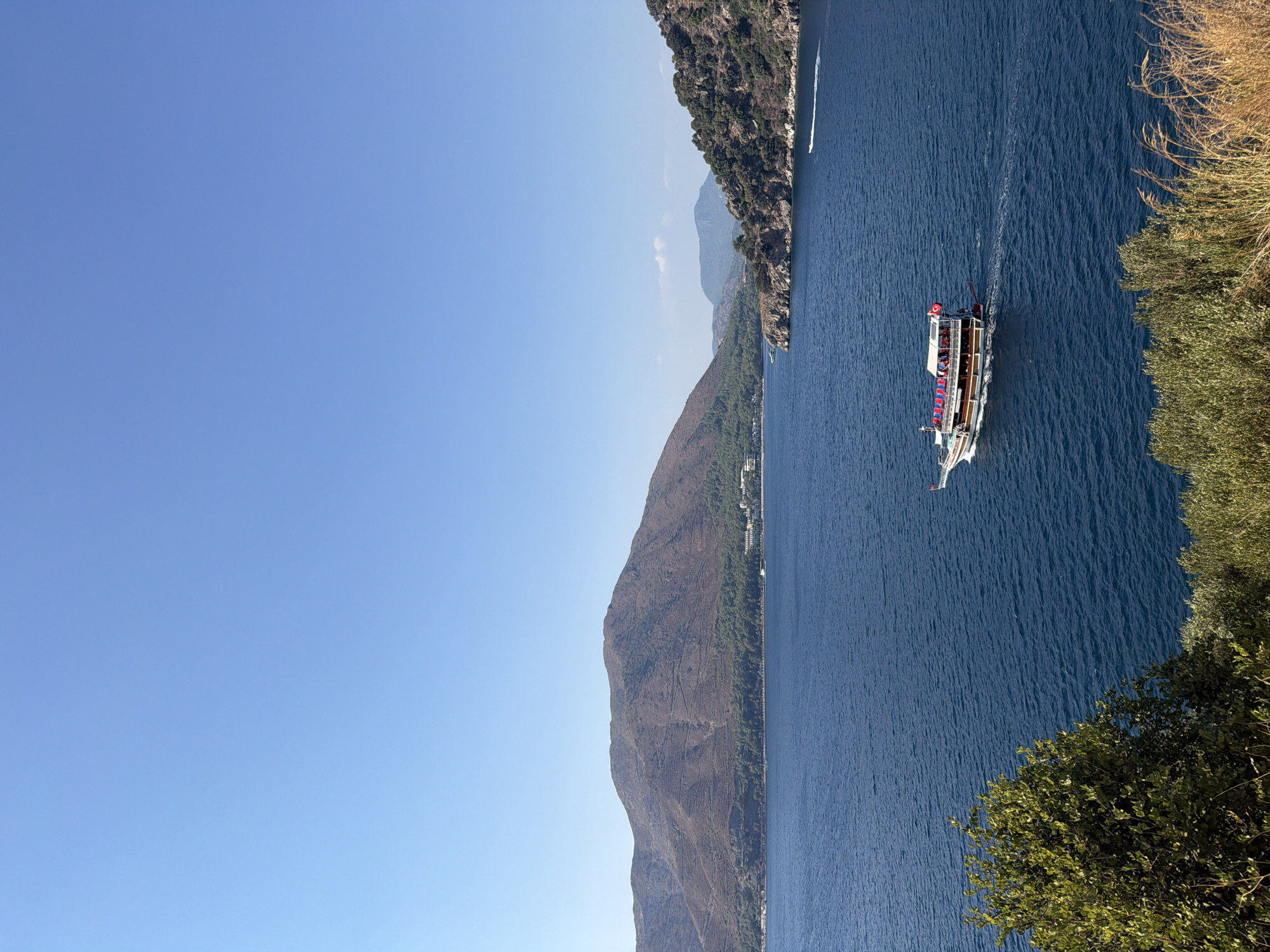 A wide, bright blue sky dominates the upper portion of the image. Below, a vast expanse of deep blue water fills the middle and lower sections. A large, multi-decked boat, decorated with red flags and awnings, cruises across the water from left to right, carrying numerous passengers. The boat has a white superstructure with brown trim. In the distance, a mountainous coastline is visible. The mountains are covered in dry, brown vegetation, with a strip of dense green trees along the shoreline. Small buildings are nestled amongst the trees on the coast. To the far right, a smaller, white boat is leaving a white wake. The foreground features a lush, green tree on the left and dry, tall grasses on the right, suggesting a natural overlook. The scene captures a sunny day with clear visibility in Marmaris, Turkey.