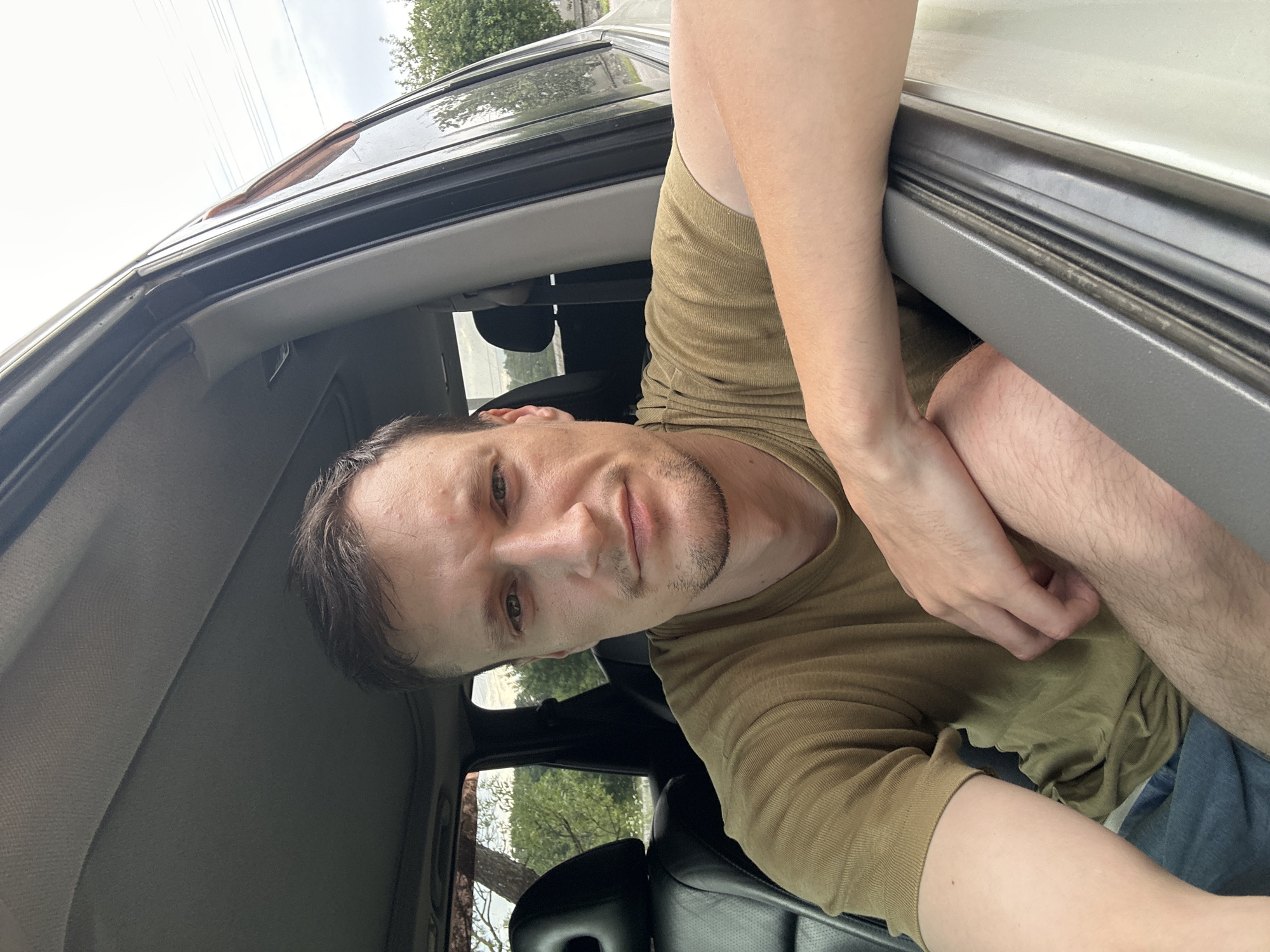 A close-up, eye-level, outdoor selfie shows a fair-skinned man with short brown hair and a slight beard, wearing a short-sleeved olive green t-shirt. He is sitting in the driver's seat of a car with the window rolled down, resting his right arm on the door frame. The background shows trees and foliage, suggesting a rural or suburban setting, consistent with the location of Makiv, Ukraine. The man is looking directly at the camera with a neutral expression. The interior of the car is partially visible, including a black leather seat and the car's headliner. The sky is overcast.