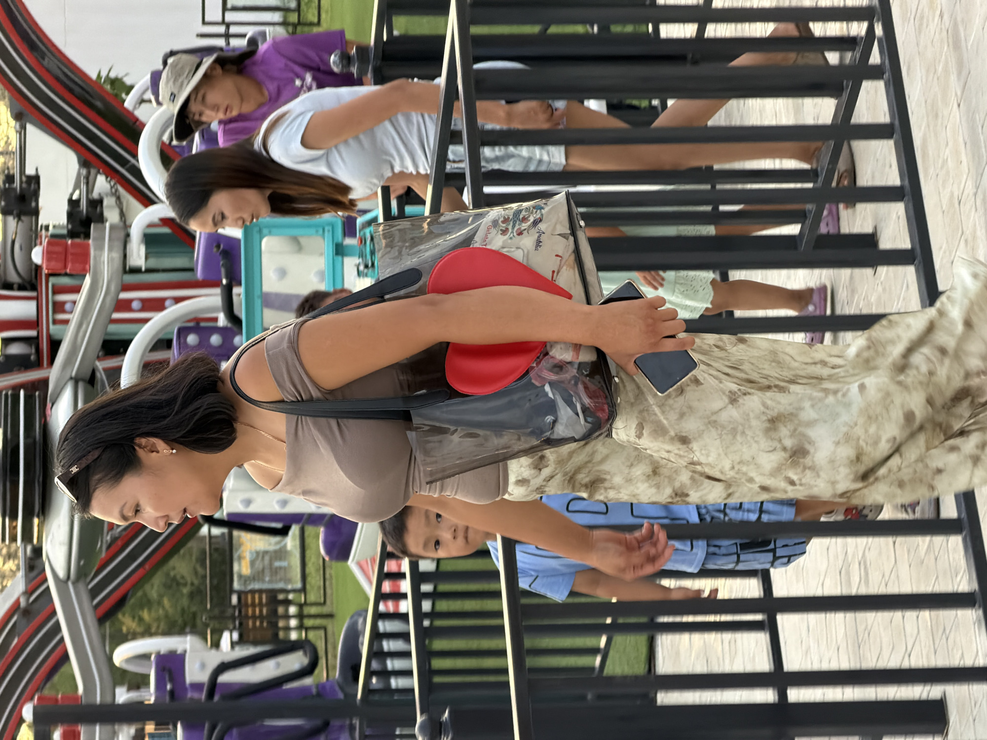An outdoor amusement park is depicted, with a group of people visible through a black metal fence. In the foreground, a woman walks to the left, wearing a beige off-the-shoulder top, a patterned skirt, and sunglasses. She carries a large transparent tote bag with a red heart-shaped item inside. A young boy in a blue shirt and blue shorts stands behind the fence to her left, looking towards the camera. To the woman's right, another woman walks away from the camera, wearing a light blue top and shorts. In the background, a young person wearing a purple shirt and a tan hat is also visible. An amusement ride with red and white striped elements is seen in the upper portion of the image.