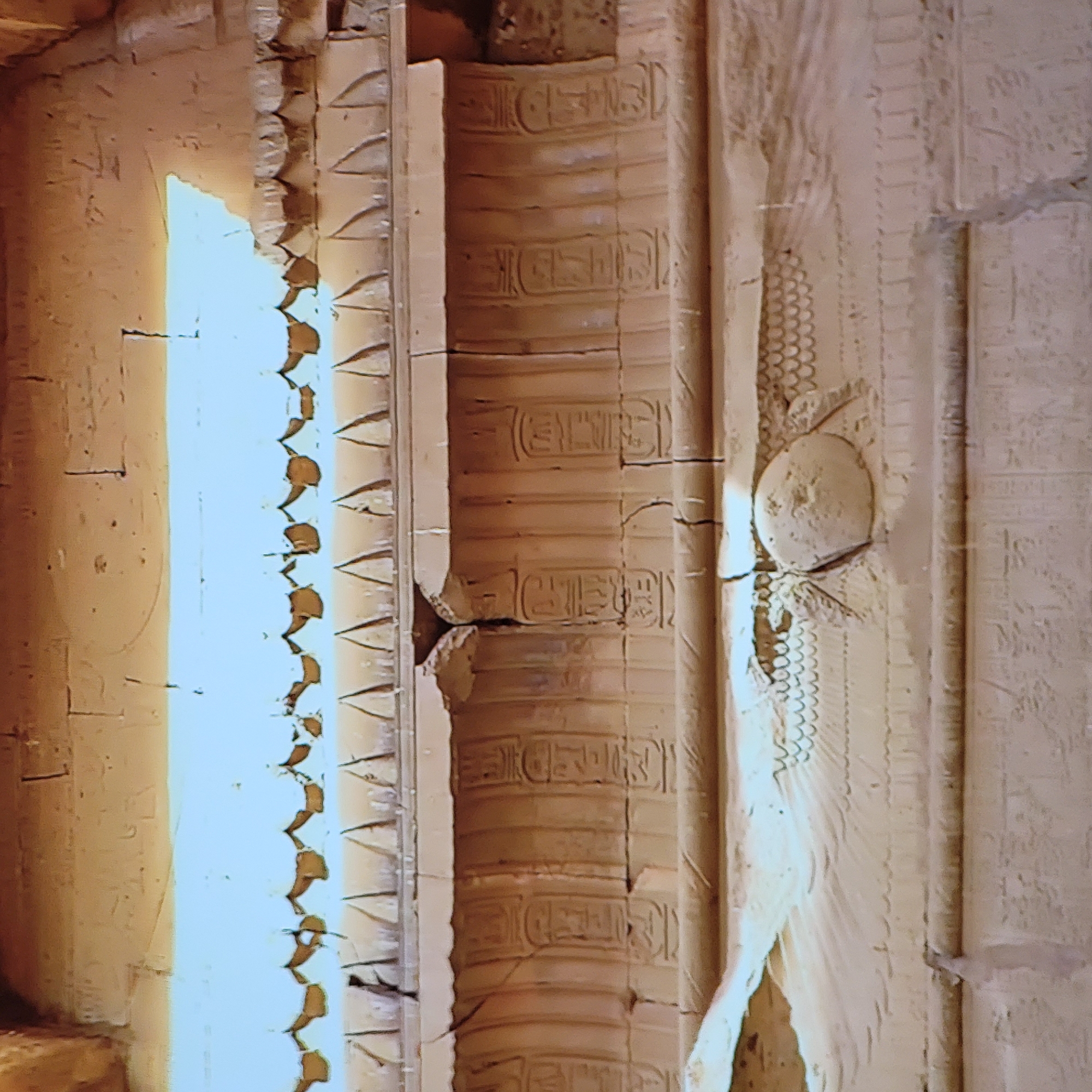 The image shows a close-up of carved stone walls, likely within an ancient Egyptian structure. Hieroglyphs are visible in vertical columns, along with decorative carvings, suggesting the walls are part of a temple or tomb. A winged sun disc is visible, indicating a religious or symbolic element. The color of the stone is a light beige or tan. The scene appears to be indoors with a light source illuminating the left side of the frame. There is no evidence to support that the location is in the city of Yerevan, Armenia.