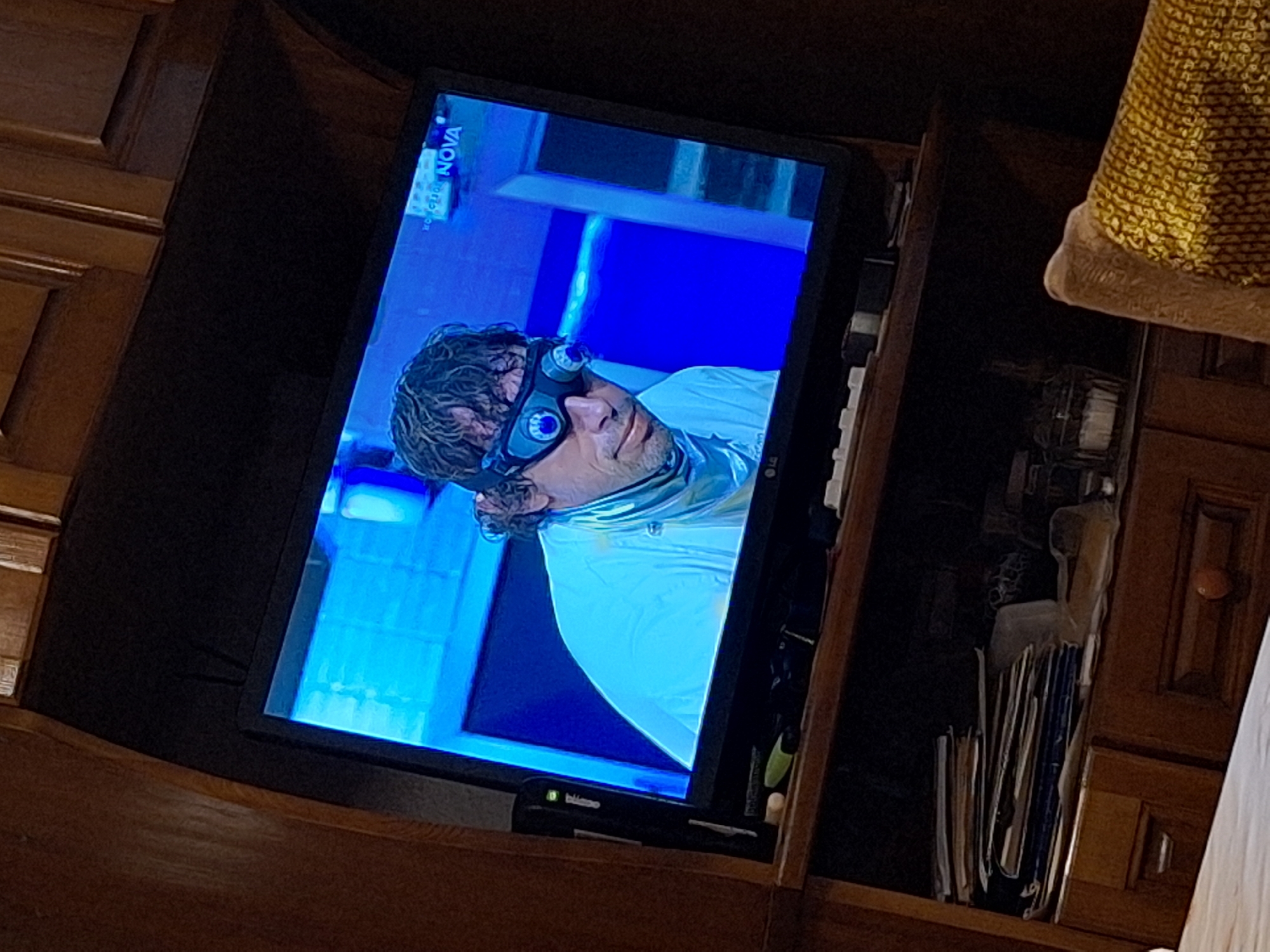 The image shows a television screen displaying a man with curly hair and goggles on his forehead. The background on the screen is blue and white. The television is set within a wooden cabinet or shelving unit, with various items such as papers and other objects stored around it. The location is unknown.