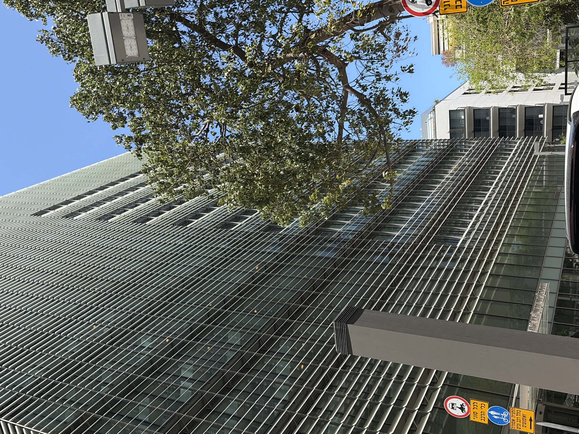 The image shows a modern building with a metal facade, likely an office building, in an urban environment. A tree with dense foliage is partially obscuring the building. Street signs in Hebrew are visible at the bottom right, indicating a location in Israel. The building's design features a grid-like pattern, with repetitive horizontal elements suggesting sunshades or architectural details. The sky is clear and blue. The location appears to be in Tel Aviv, Israel, as indicated by the Hebrew street signs.
